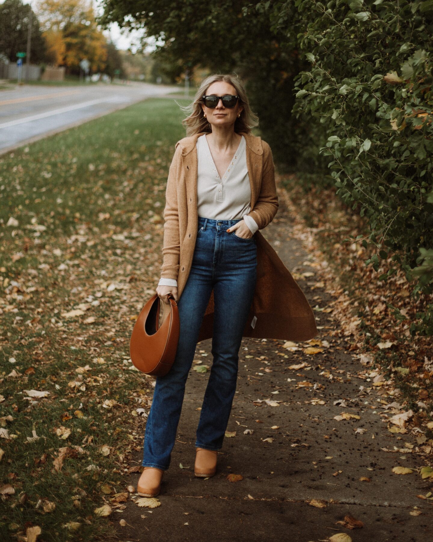 Karin Emily wears a henley tee, a long maxi cardigan from Everlane, boot leg sustainable jeans from Revolve, clogs, and a staud moon bag