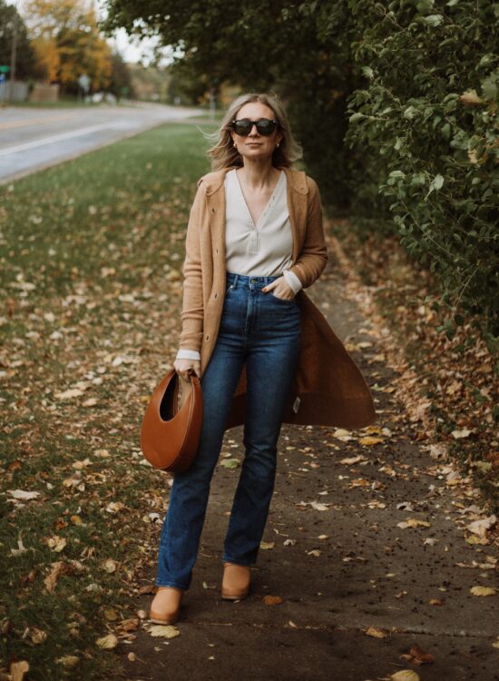 Karin Emily wears a henley tee, a long maxi cardigan from Everlane, boot leg sustainable jeans from Revolve, clogs, and a staud moon bag