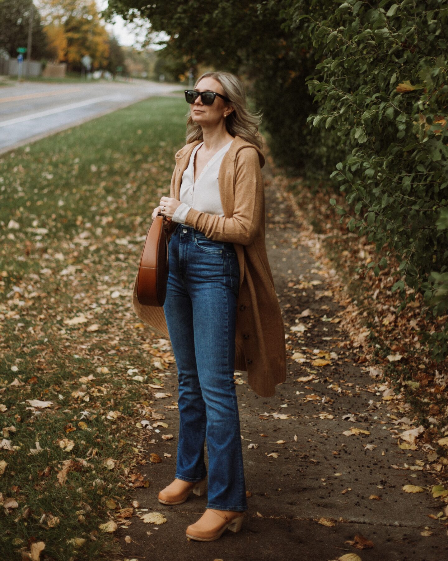 Karin Emily wears a henley tee, a long maxi cardigan from Everlane, boot leg sustainable jeans from Revolve, clogs, and a staud moon bag