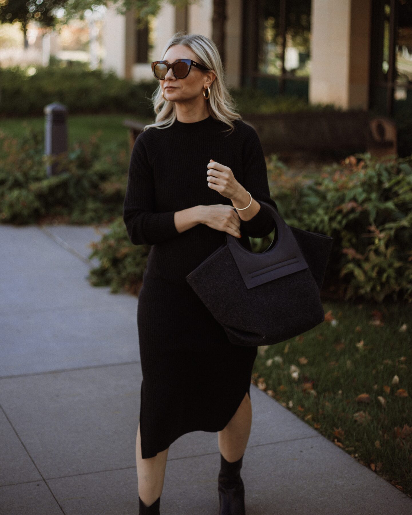 Karin Emily wears a black sweater dress, hereu bag, black cowboy boots, and oversized black sunglasses