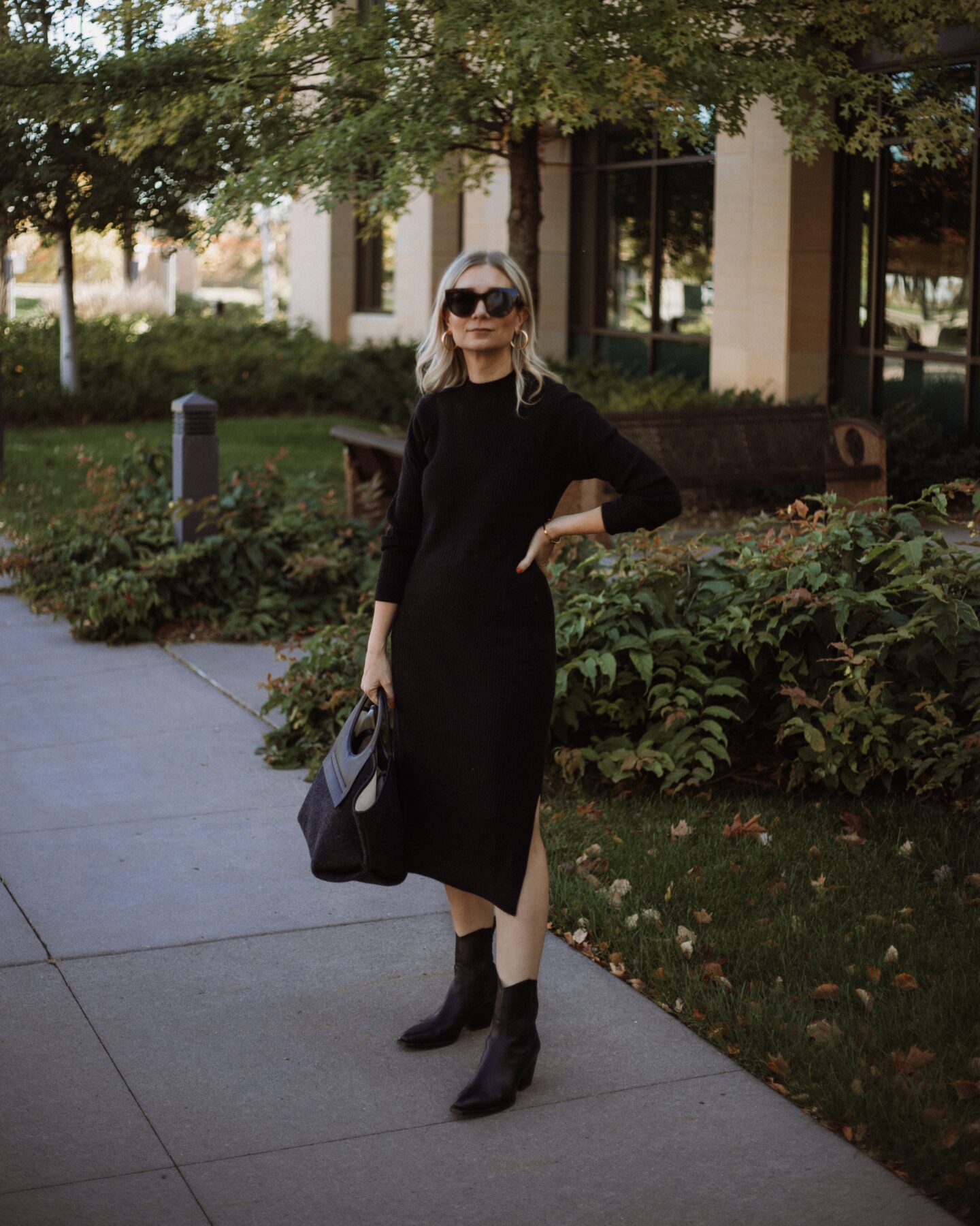 Karin Emily wears a black sweater dress, hereu bag, black cowboy boots, and oversized black sunglasses