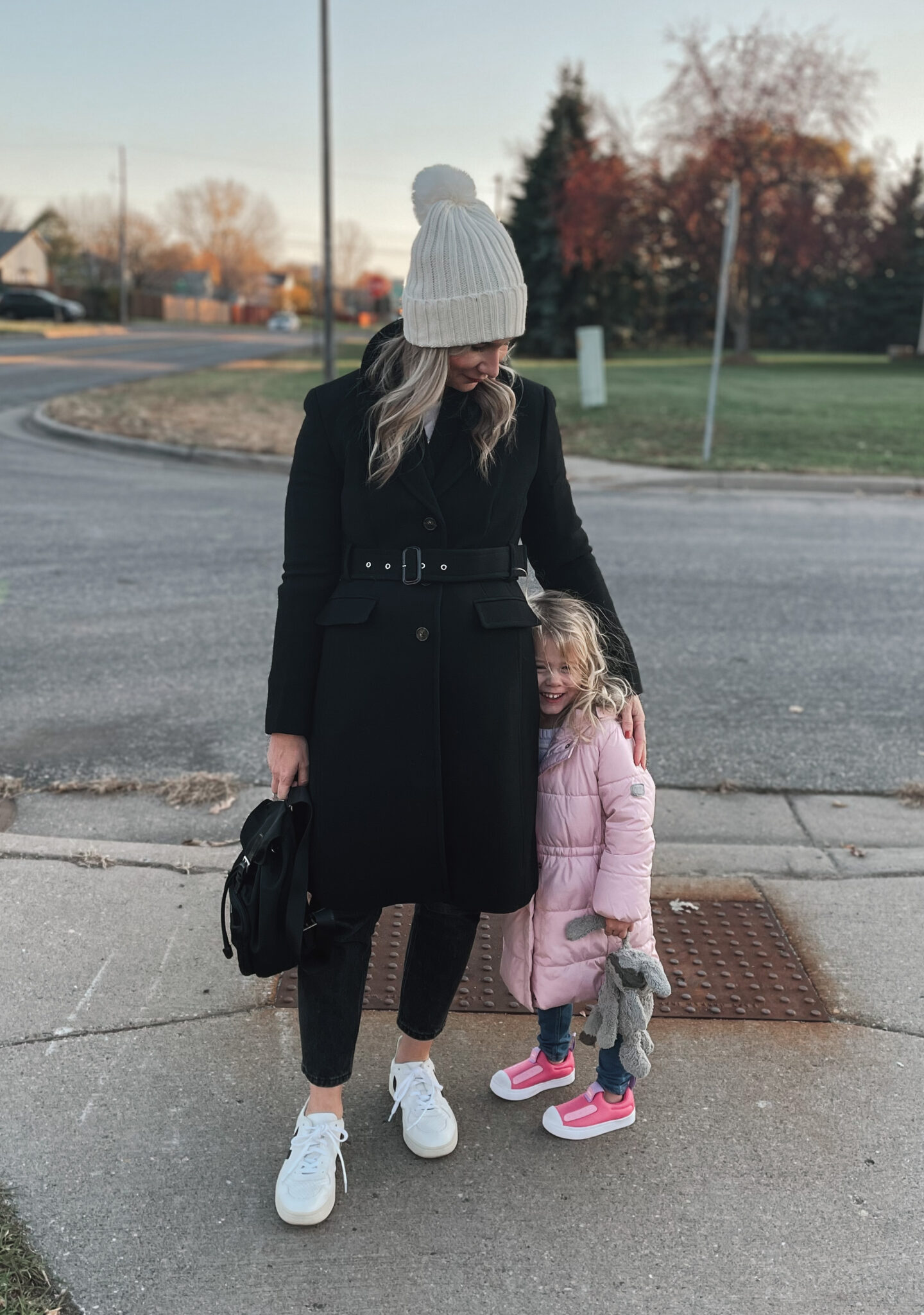 Karin Emily wears a pom snow hat, Black belted wool coat, black jeans, and white Veja V-10 sneakers