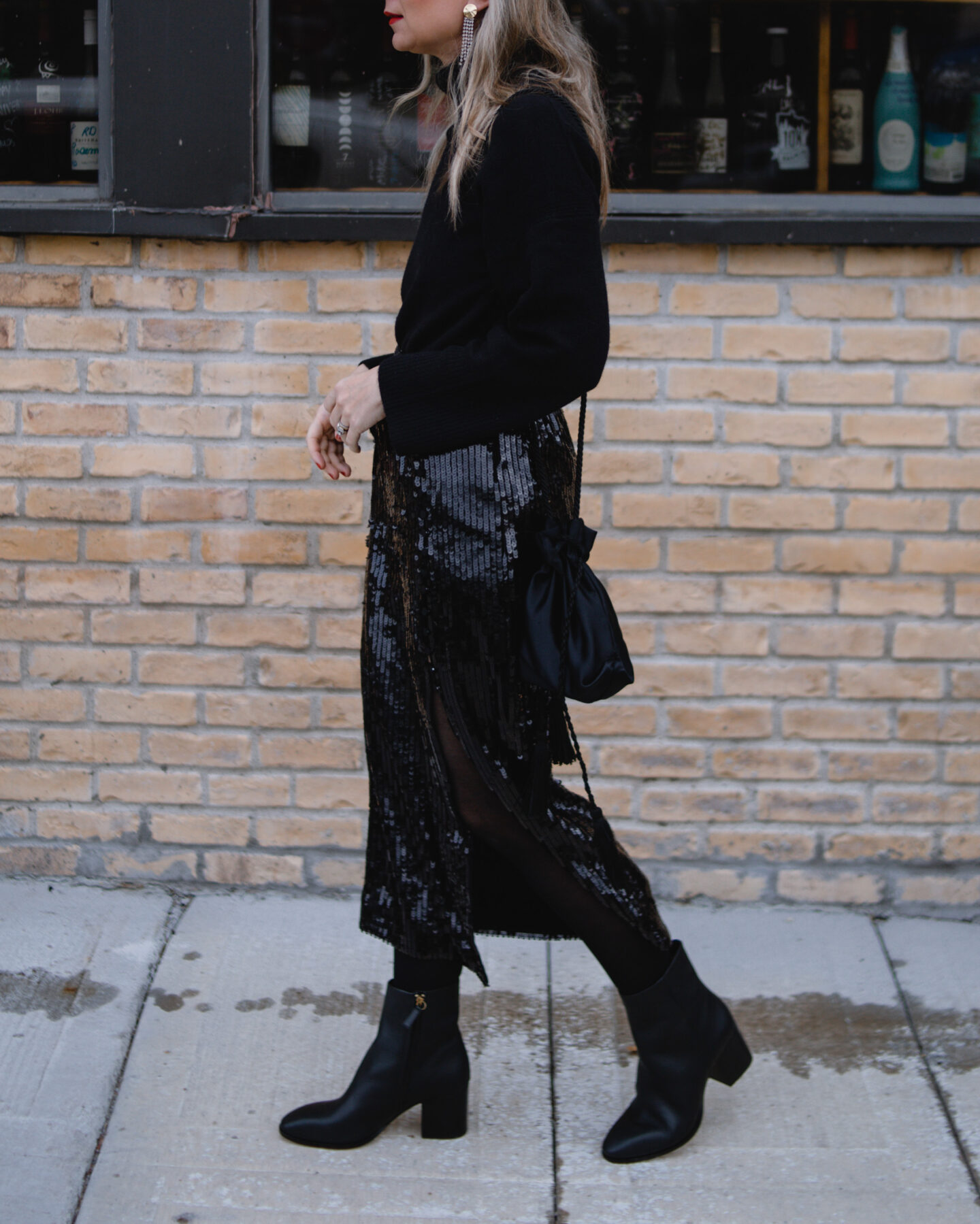 Karin Emily wears a black turtleneck sweater, sequin skirt, and black heeled booties with a pair of cat eye sunglasses