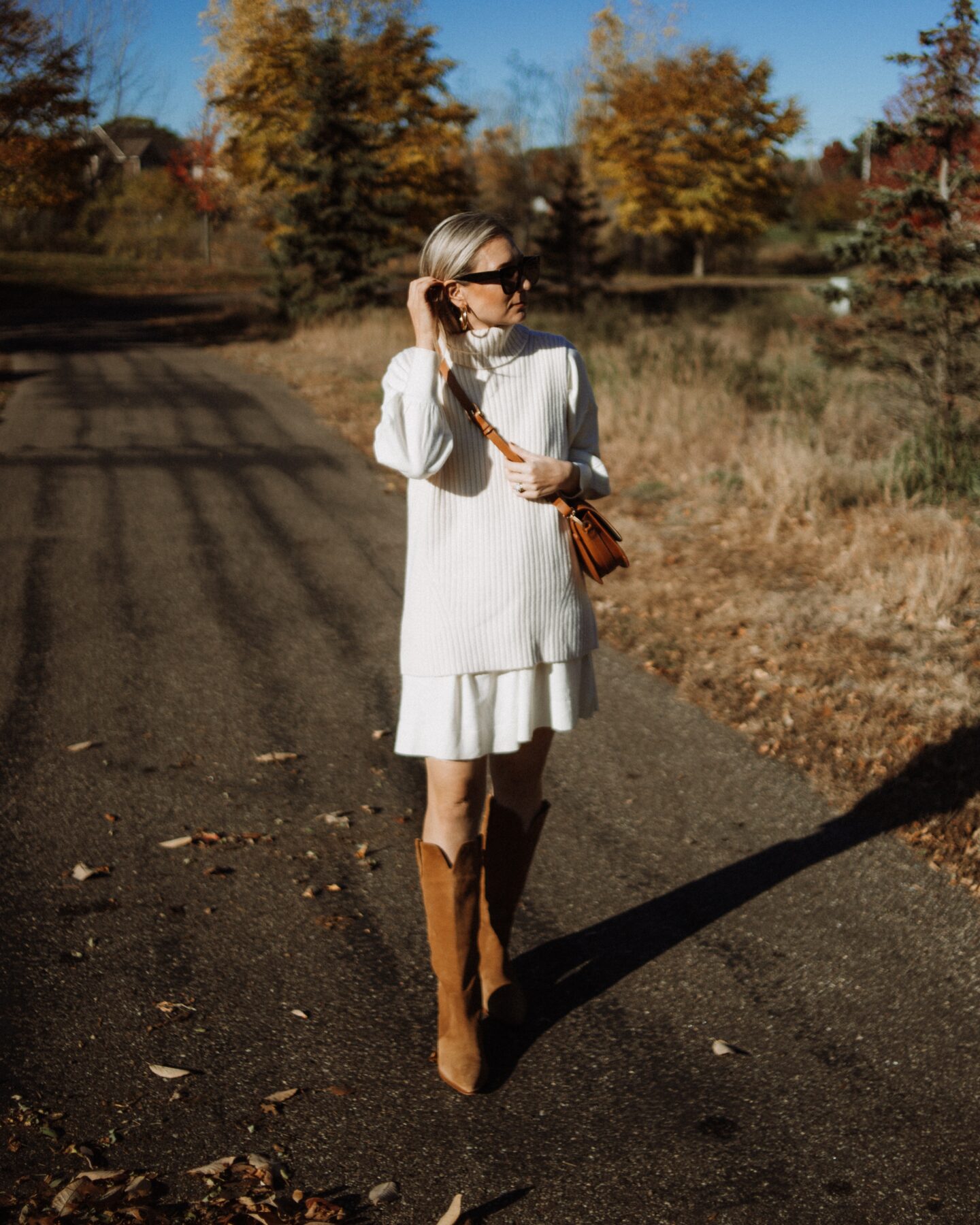 Karin Emily wears a cream sweater vest over a cream sweater dress and knee high western boots with a brown saddle bag