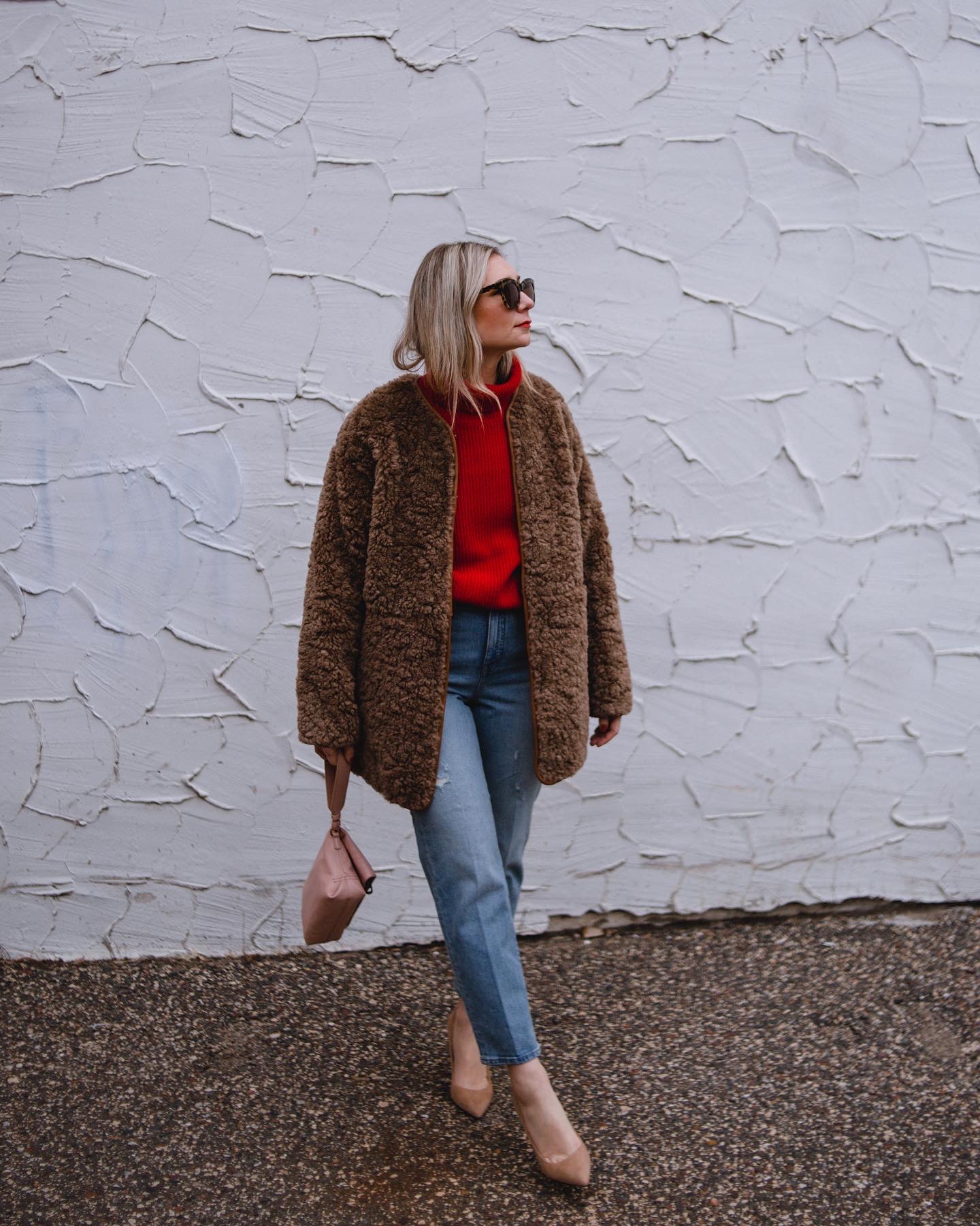 Karin Emily wears a brown sherpa coat, red cashmere sweater, blue 90's jeans, and suede nude heels