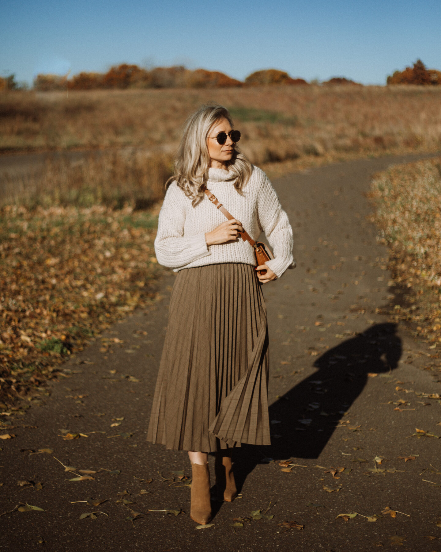 Karin Emily wears a cropped sweater from the Caitlin Covington for Pink Lily Collection, a Pleated Skirt, a Brown Saddle Bag, and a pair of Marc Fisher Brown Booties