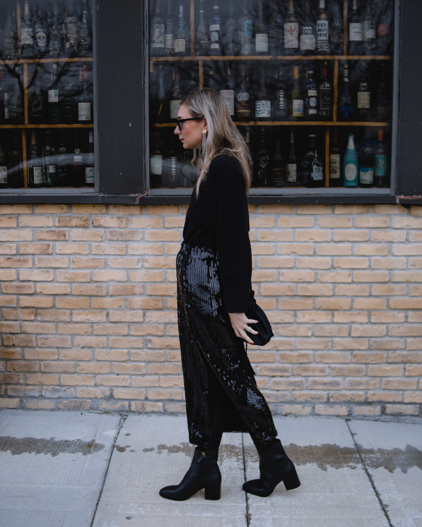 Karin Emily wears a black turtleneck sweater, sequin skirt, and black heeled booties with a pair of cat eye sunglasses