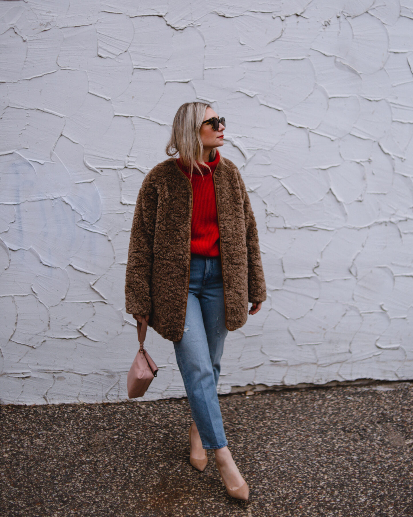 Karin Emily wears a red cashmere sweater, camel colored sherpa coat, and mid wash straight leg jeans, and nude suede heels