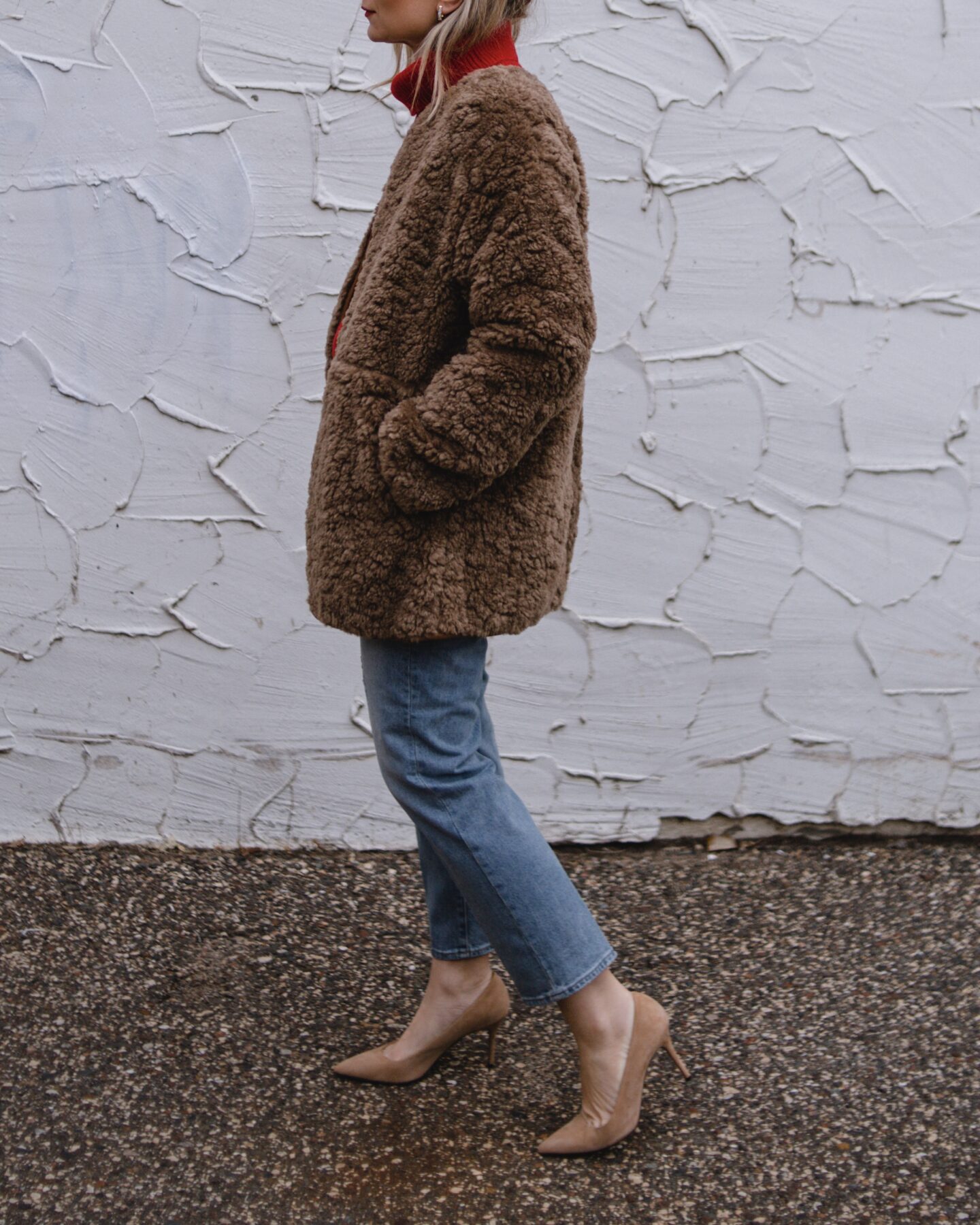 Karin Emily wears a red cashmere sweater, camel colored sherpa coat, and mid wash straight leg jeans, and nude suede heels