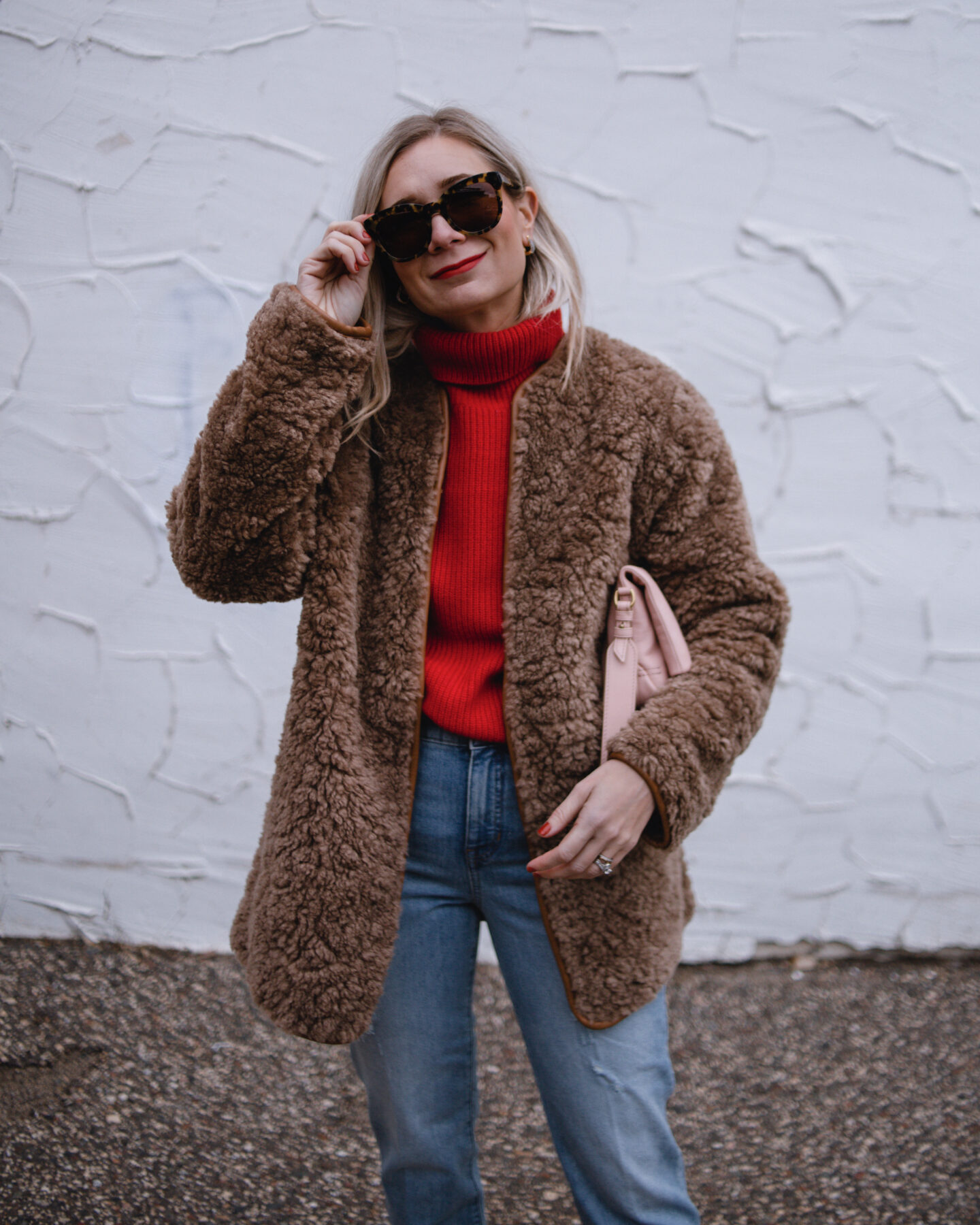 Karin Emily wears a red cashmere sweater, camel colored sherpa coat, and mid wash straight leg jeans