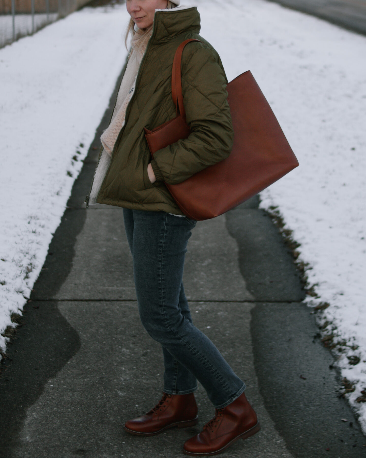 Karin Emily wears an olive green puffer coat, dark wash straight leg jeans, a brown tote bag, and brown lace up boots