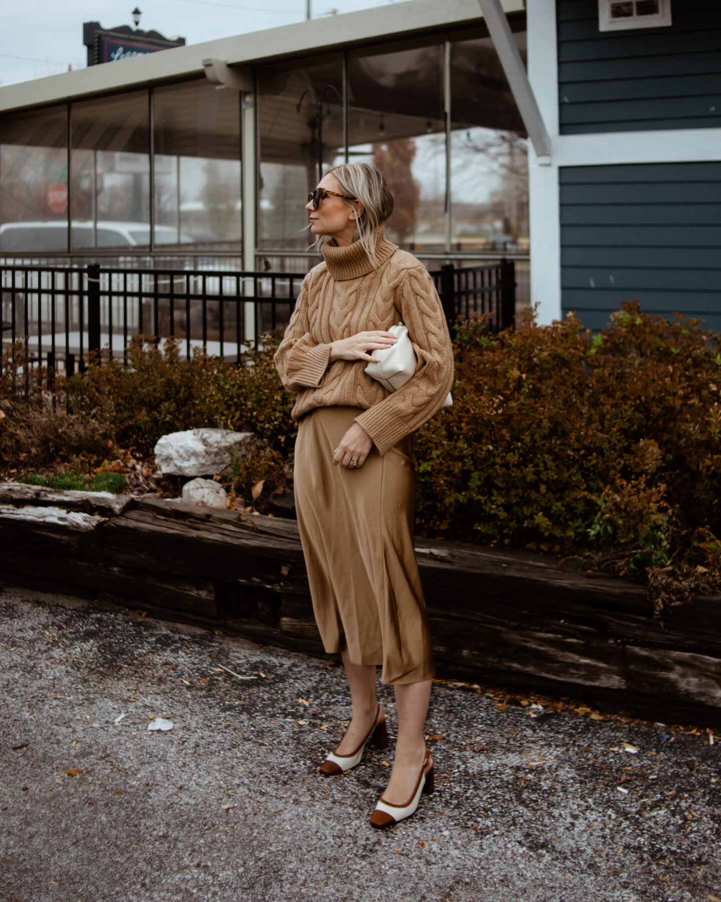 Karin Emily wears a camel cable knit sweater, satin slip skirt, and cap toe heels