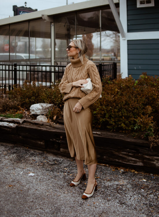 Karin Emily wears a camel cable knit sweater, satin slip skirt, and cap toe heels
