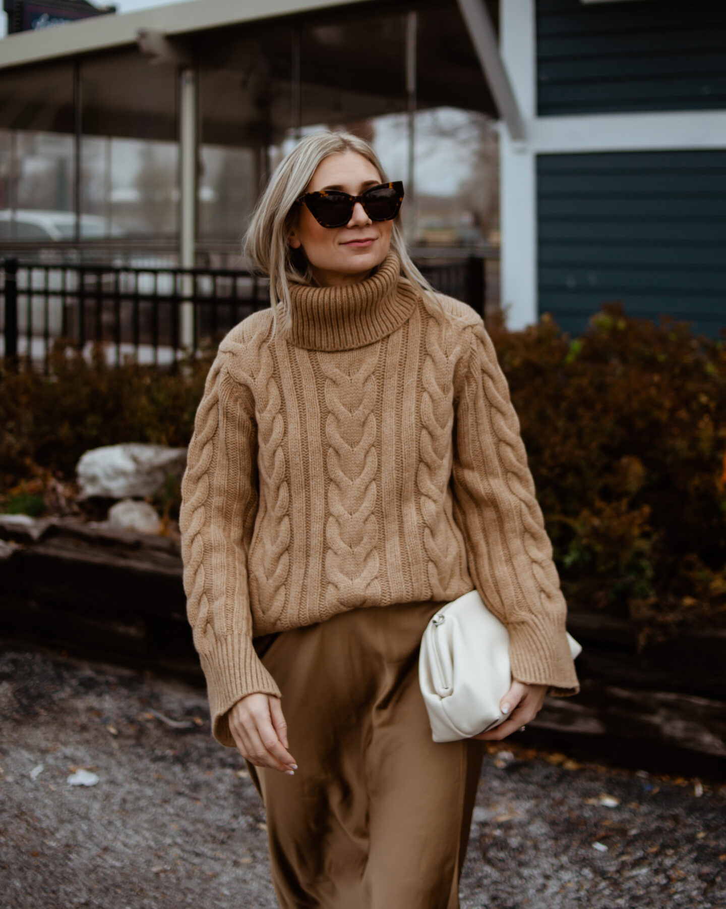 Karin Emily wears a camel cable knit sweater, satin slip skirt, and a cream clutch