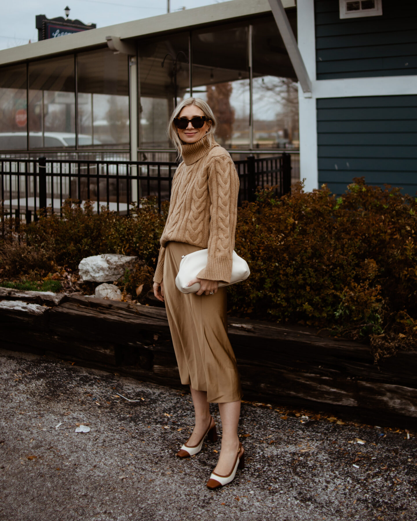 Karin Emily wears a camel cable knit sweater, satin slip skirt, and cap toe heels