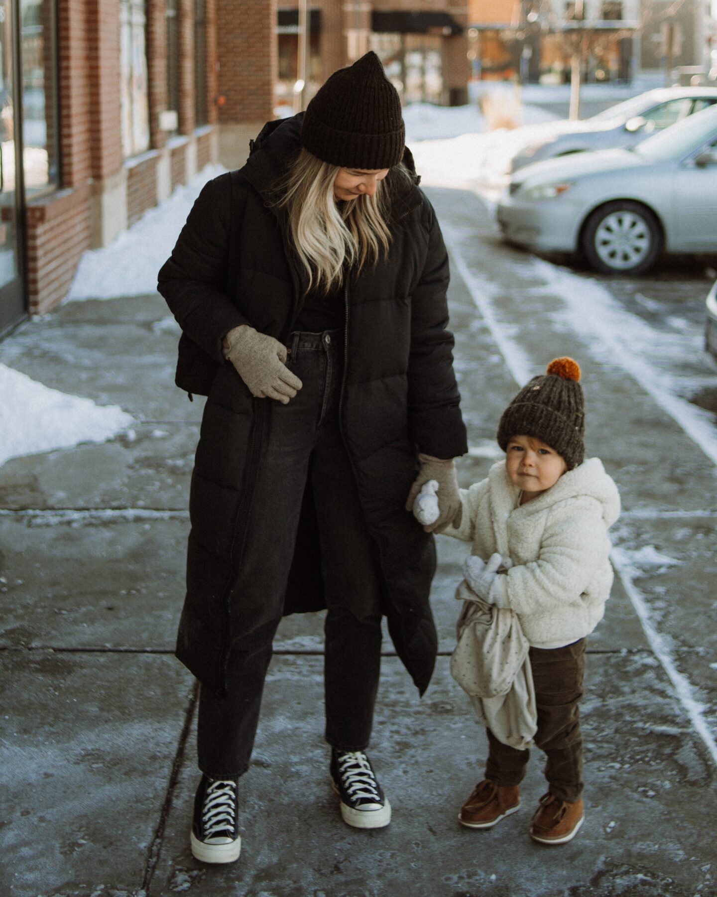 Karin Emily stands with her toddler and wears a black maxi puffer coat from Girlfriend, a ribbed black long sleeve tee from Madewell, Agolde 90's jeans, and black high top converse 