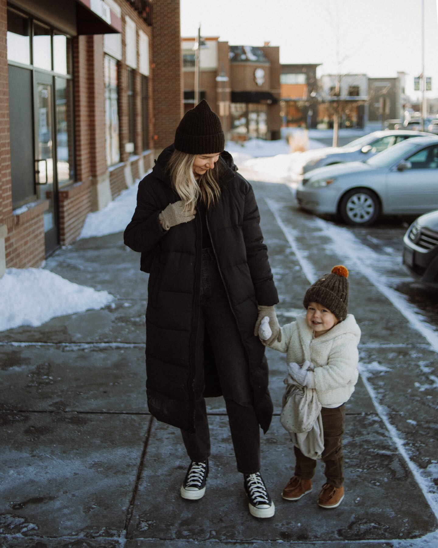 Karin Emily stands with her toddler and wears a black maxi puffer coat from Girlfriend, a ribbed black long sleeve tee from Madewell, Agolde 90's jeans, and black high top converse