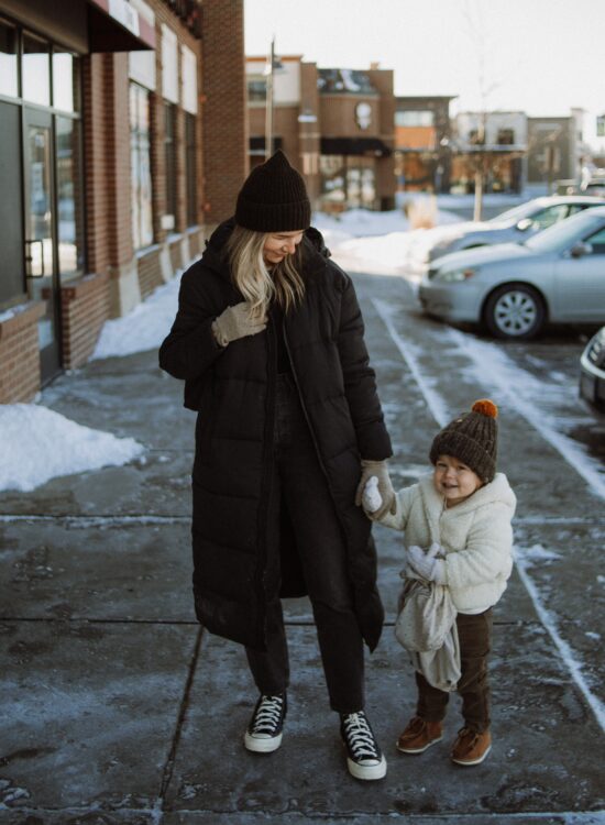 Karin Emily stands with her toddler and wears a black maxi puffer coat from Girlfriend, a ribbed black long sleeve tee from Madewell, Agolde 90's jeans, and black high top converse