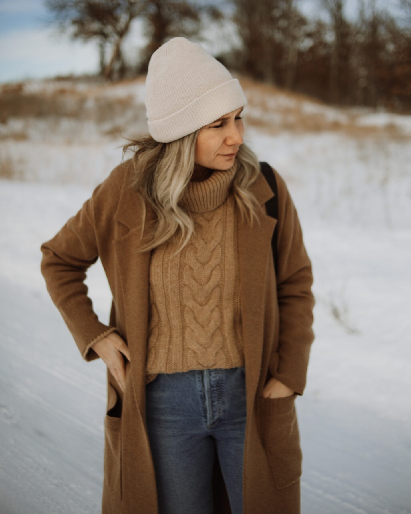 Karin Emily wears a camel sweater coat, camel chunky turtleneck sweater, agolde riley distressed jeans, and veja venturi sneakers