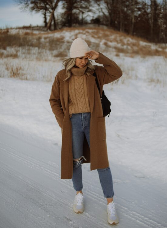 Karin Emily wears a camel sweater coat, camel chunky turtleneck sweater, agolde riley distressed jeans, and veja venturi sneakers