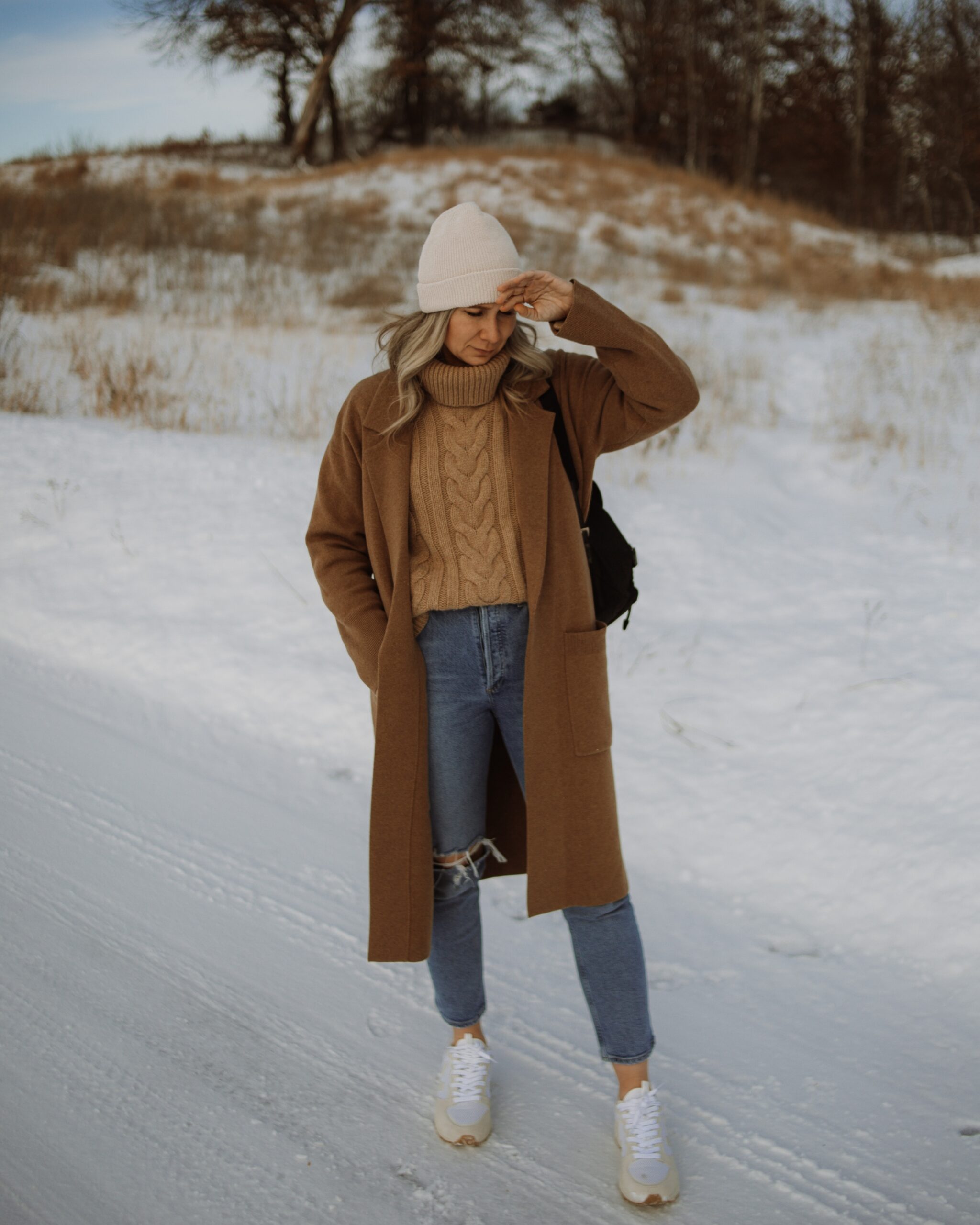Karin Emily wears a camel sweater coat, camel chunky turtleneck sweater, agolde riley distressed jeans, and veja venturi sneakers