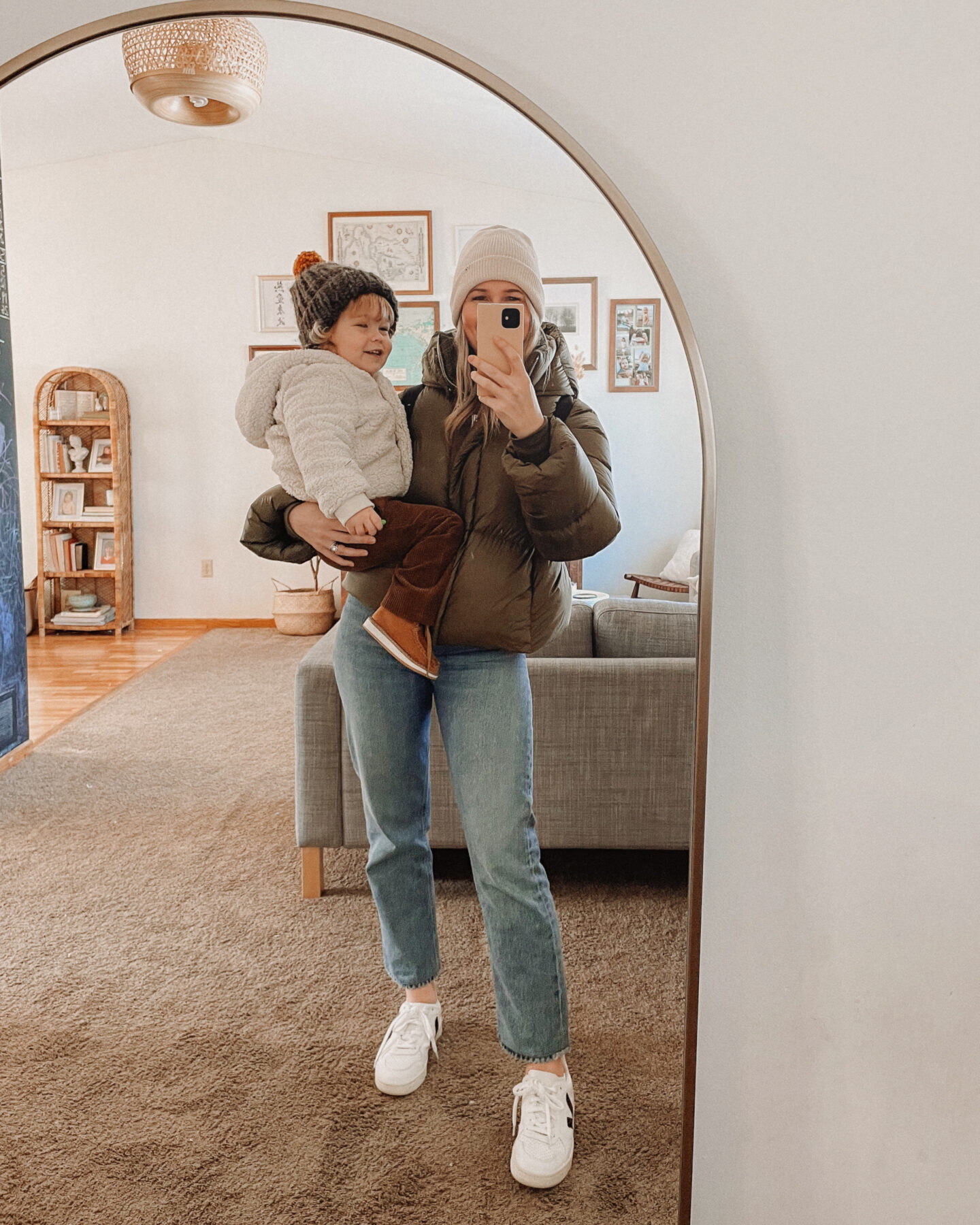 Karin Emily wears an olive green cropped puffer coat from Banana Republic, Agolde Pinch Waist Straight Leg Jeans in a blue mid wash, and Veja V-10 Sneakers while holding her son