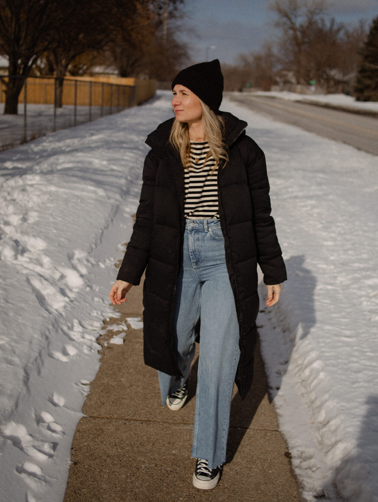 Karin Emily Wears a long black puffer coat, a striped tee, wide leg jeans, and black converse
