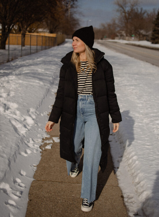 Karin Emily Wears a long black puffer coat, a striped tee, wide leg jeans, and black converse
