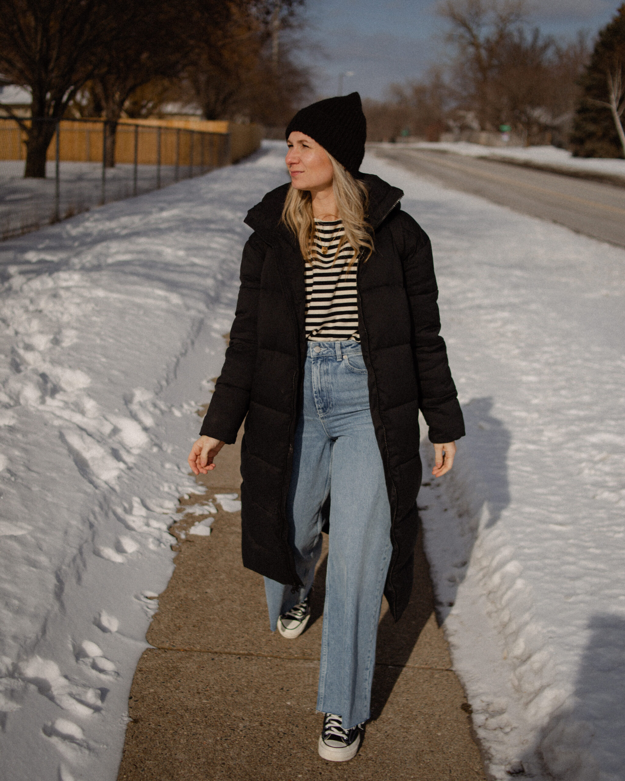 Karin Emily Wears a long black puffer coat, a striped tee, wide leg jeans, and black converse