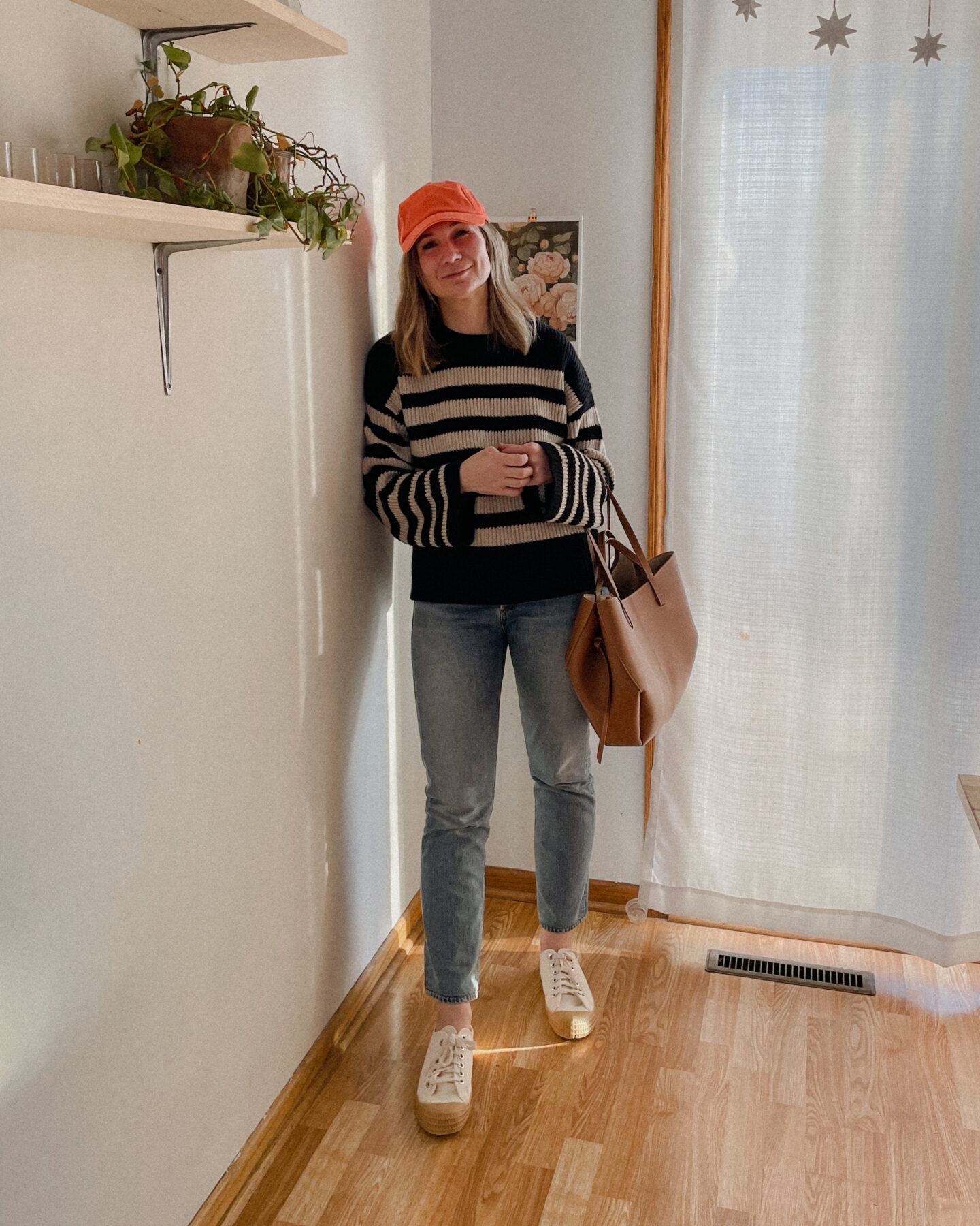 Karin Emily wears a pop of vibrant orange in the form of a baseball hat paired with a breton stripe sweater, straight leg jeans, and canvas sneakers