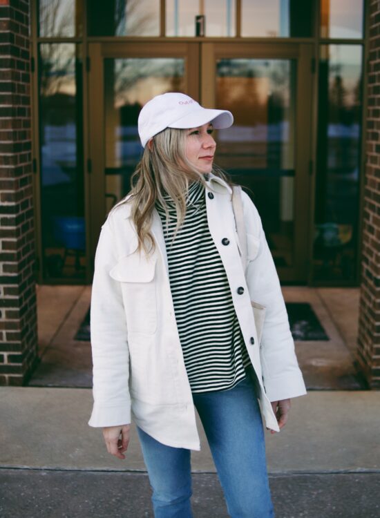 Karin Emily wears a white shirt jacket and striped sweatshirt from Everlane, blue denim Agolde jeans, and Chanel Inspired Two Tone Flats