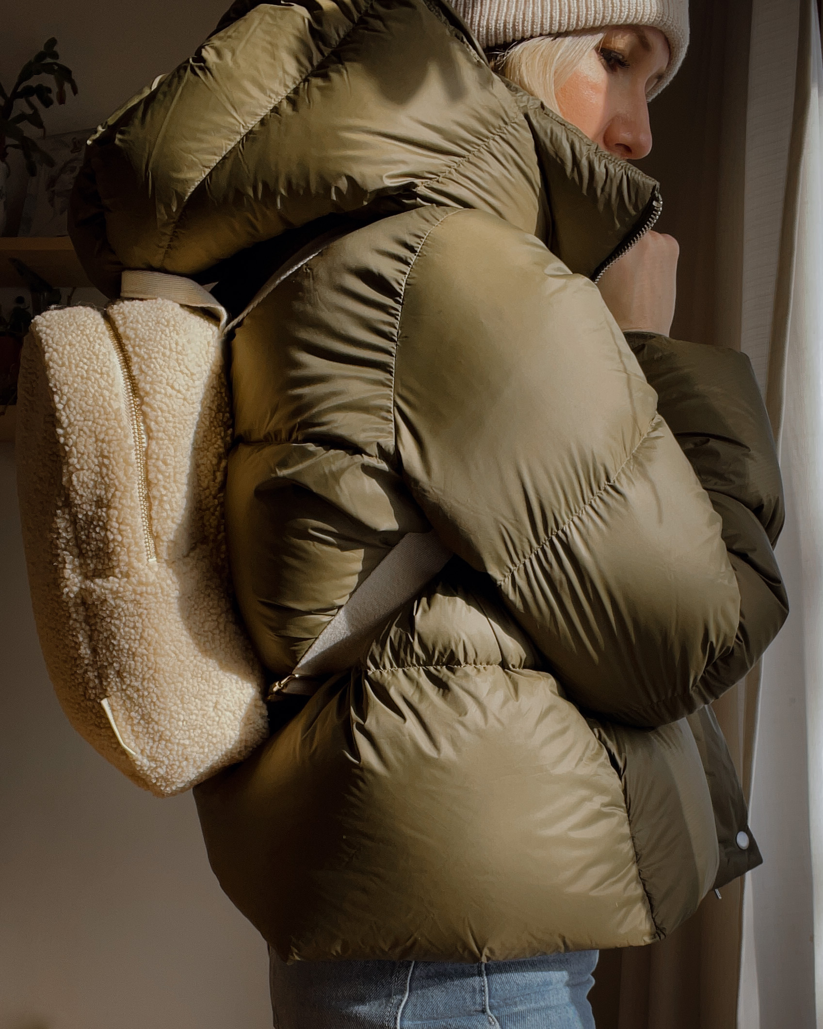 Karin Emily wears a cropped army green puffer coat, cream turtleneck sweater and sherpa backpack