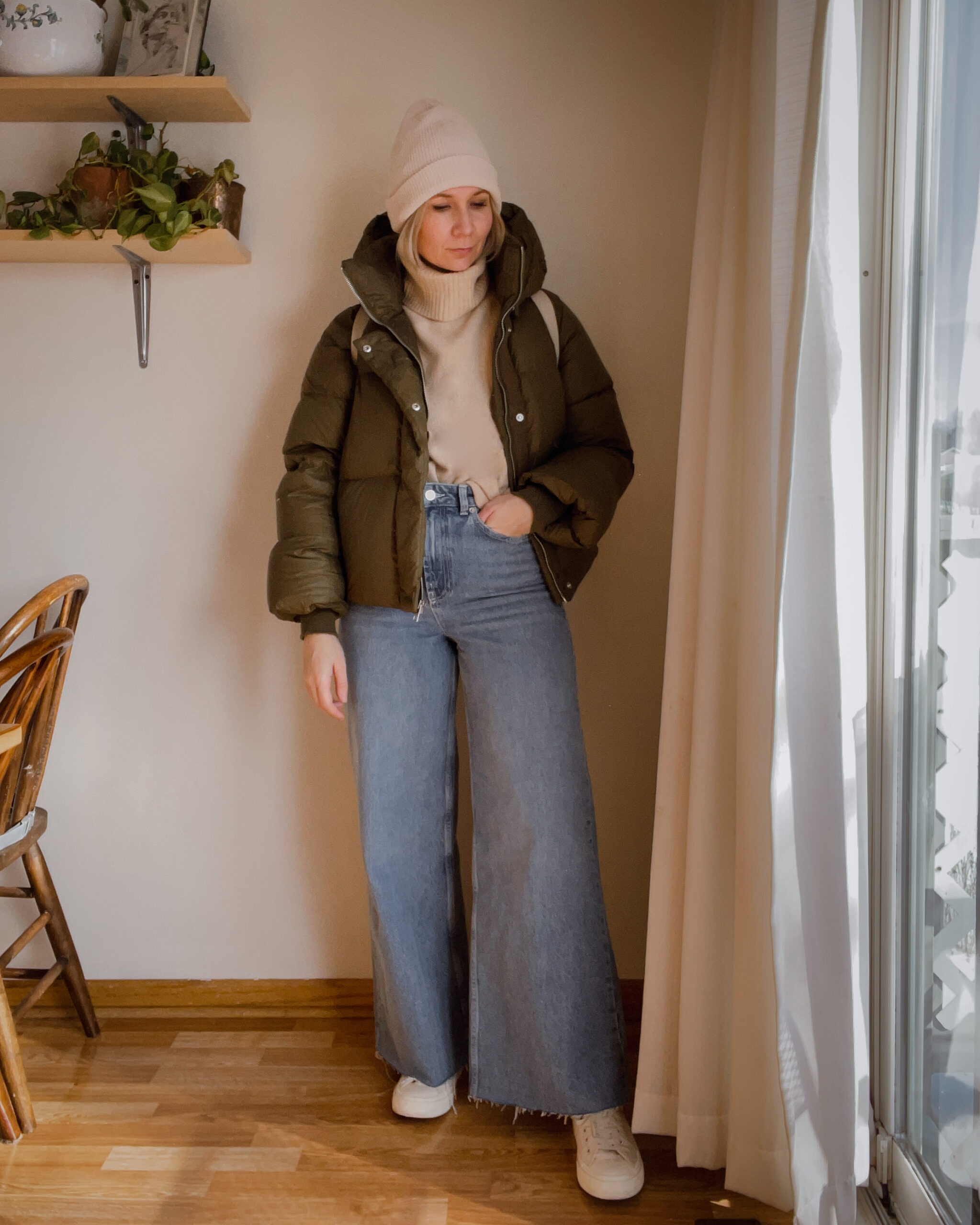Karin Emily wears a cropped army green puffer coat, cream turtleneck sweater and winter hat, wide leg jeans, and off white converse