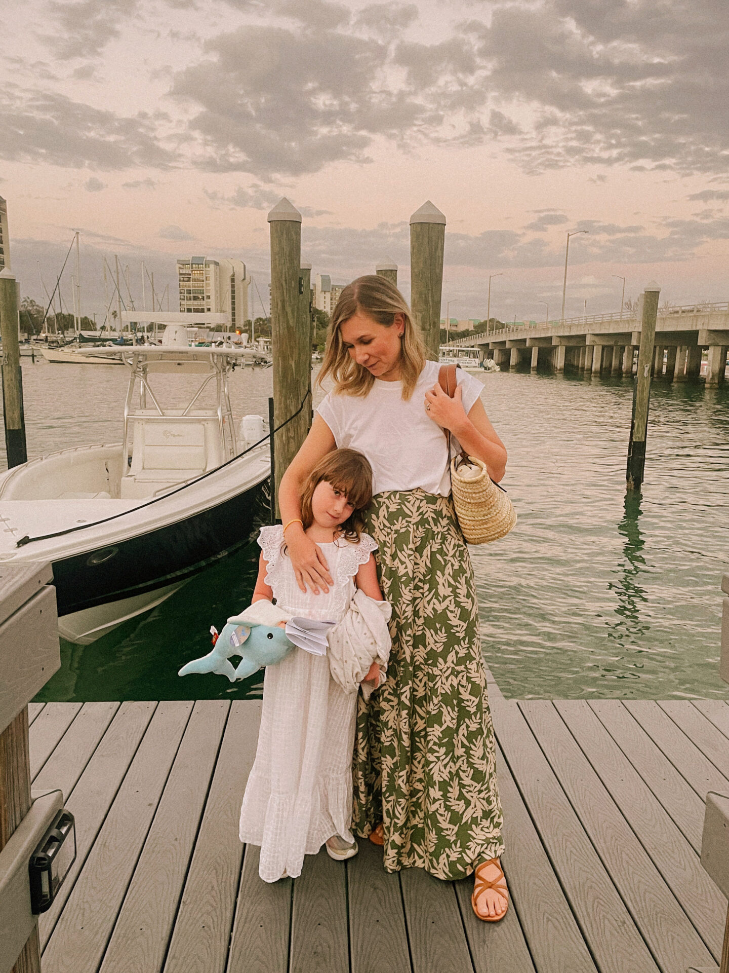 Karin Emily wears resort wear: a long green maxi skirt, a perfect white tee, and brown lace up sandals next to her daughter who's wearing a maxi length white lace dress
