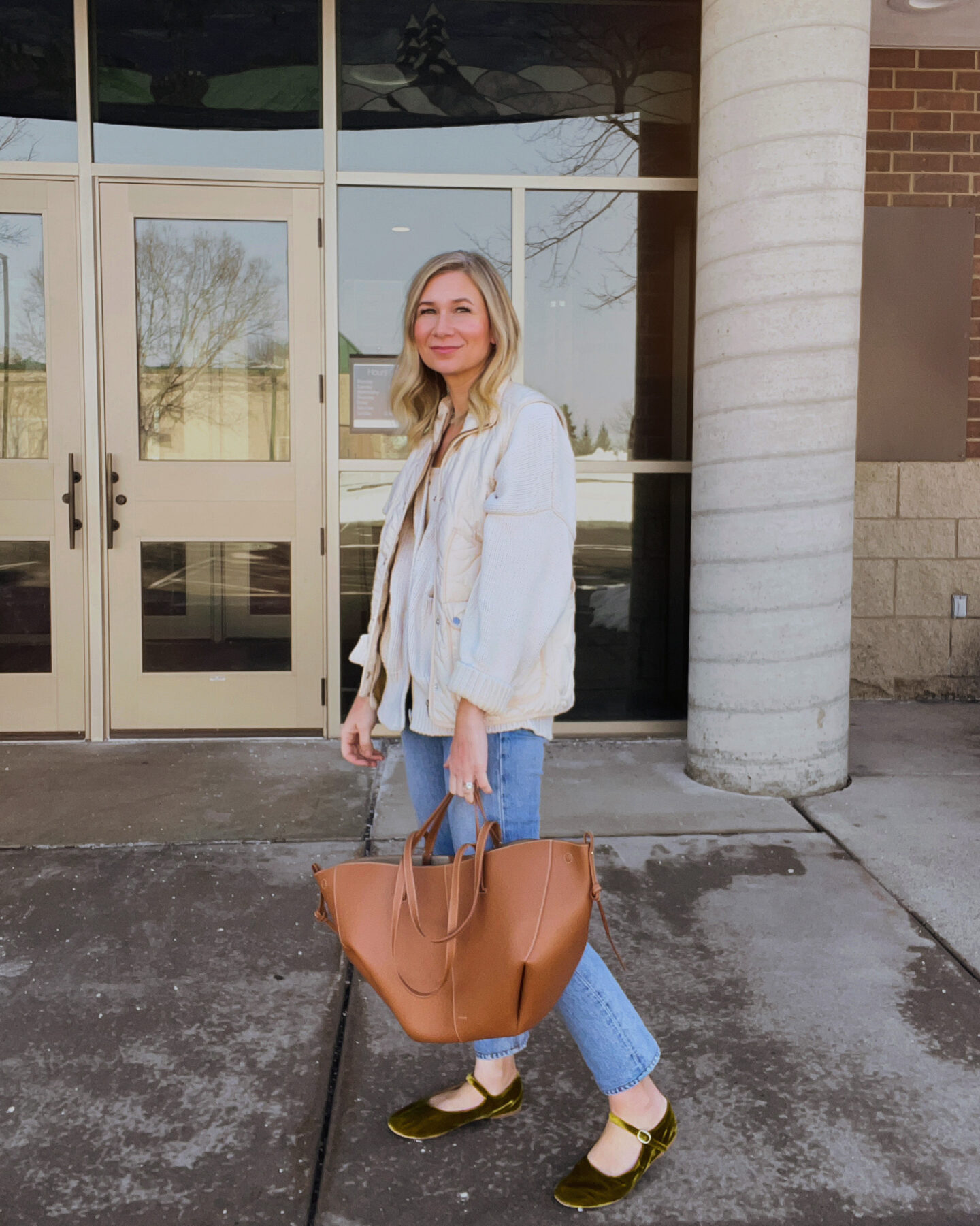 Karin Emily wears a cotton cardigan, cream vest, straight leg light wash jeans, an oversized brown tote bag, and green velvet Mary Janes