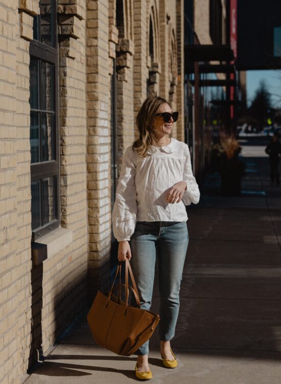 Karin Emily wears a white linen peter pan collar shirt with a pair of light wash straight leg jeans, yellow ballet flats, and an oversized polene tote bag