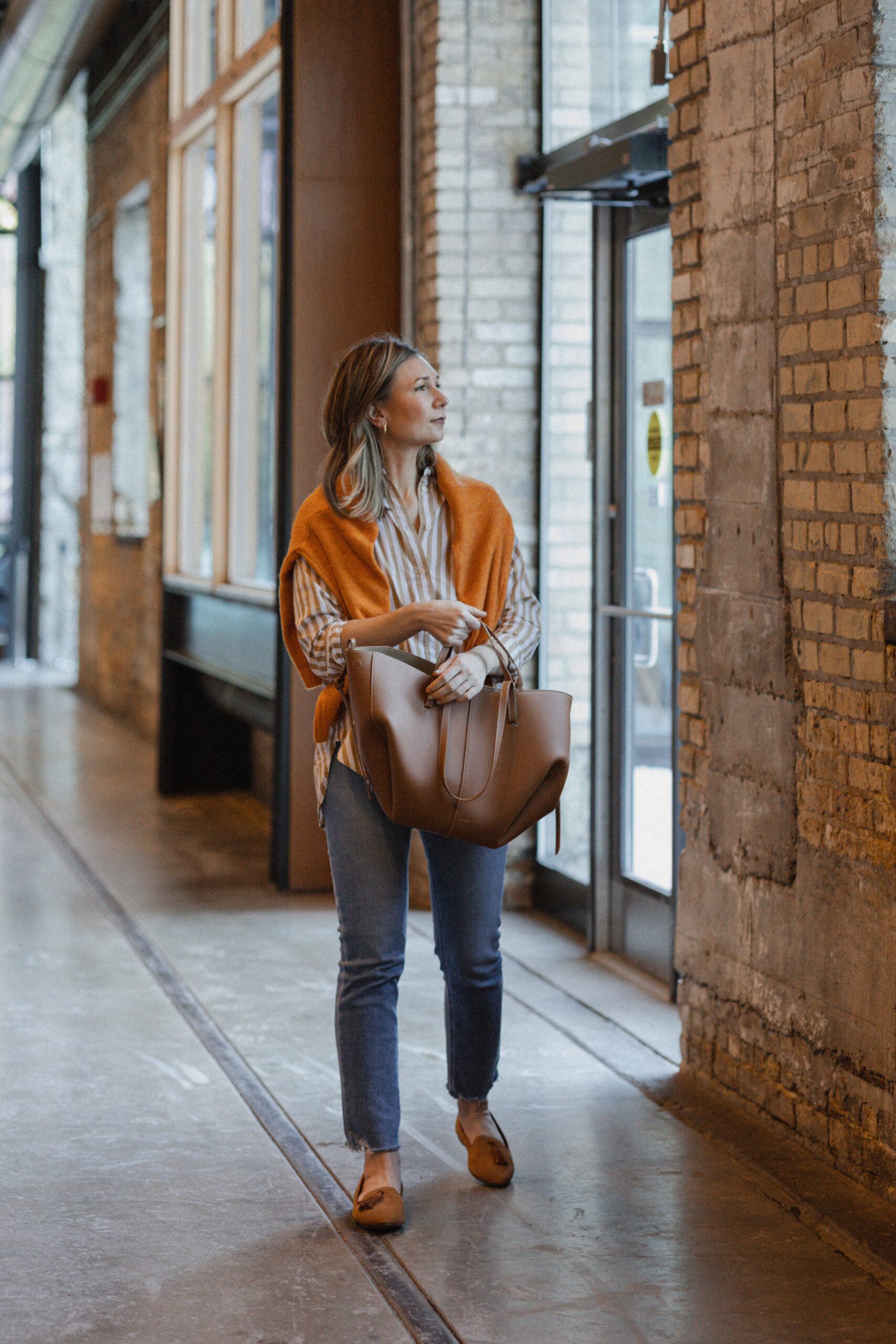 Karin Emily wears an orange stripe button down with an orange sweater over her shoulders paired with a comfy pair of jeans, loafers and an oversized tote bag