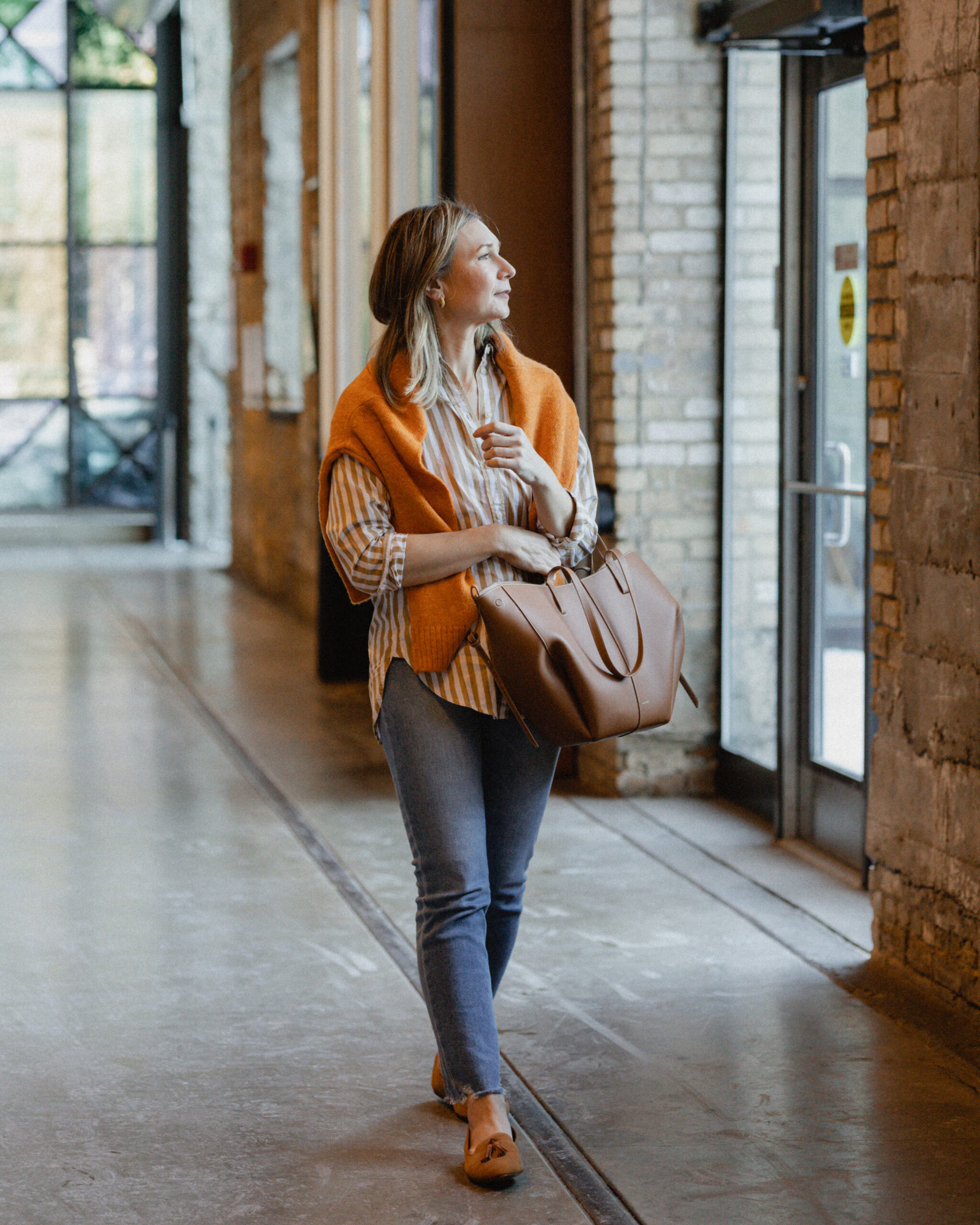 Karin Emily wears an orange stripe button down with an orange sweater over her shoulders paired with a comfy pair of jeans, loafers and an oversized tote bag
