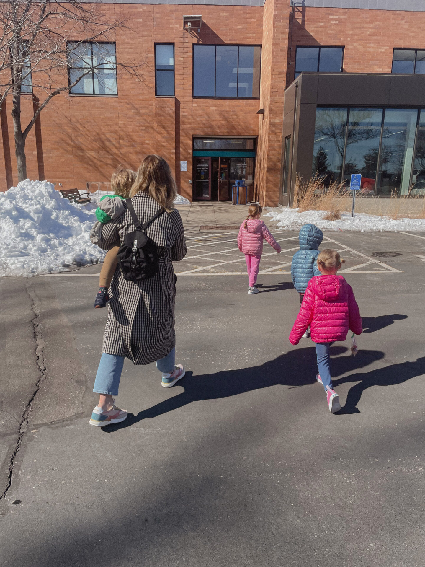 Karin Emily wears a gingham trench coat with straight leg jeans and rainbow sneakers with her kids who are wearing bright colored puffer coats