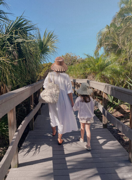 Karin Emily wears resort wear: a long white gauze beach coverup dress with a lorna murray hat next to her daughter who's wearing a lavender sweatshirt, floral shorts, and a green bucket hat