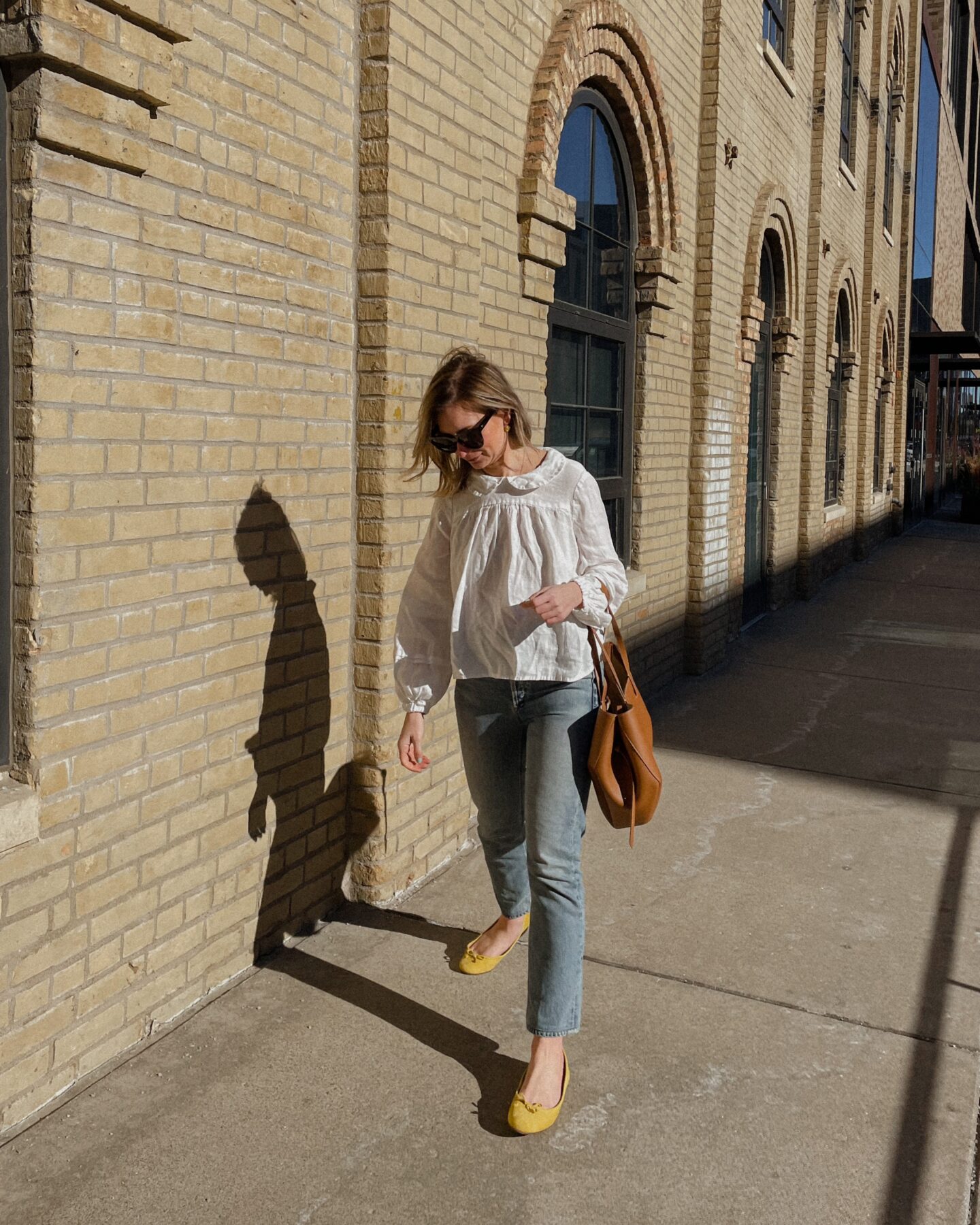 Karin Emily wears a white Peter Pan collar blouse with a pair of light wash straight leg jeans, yellow Flats, and an oversized brown tote bag