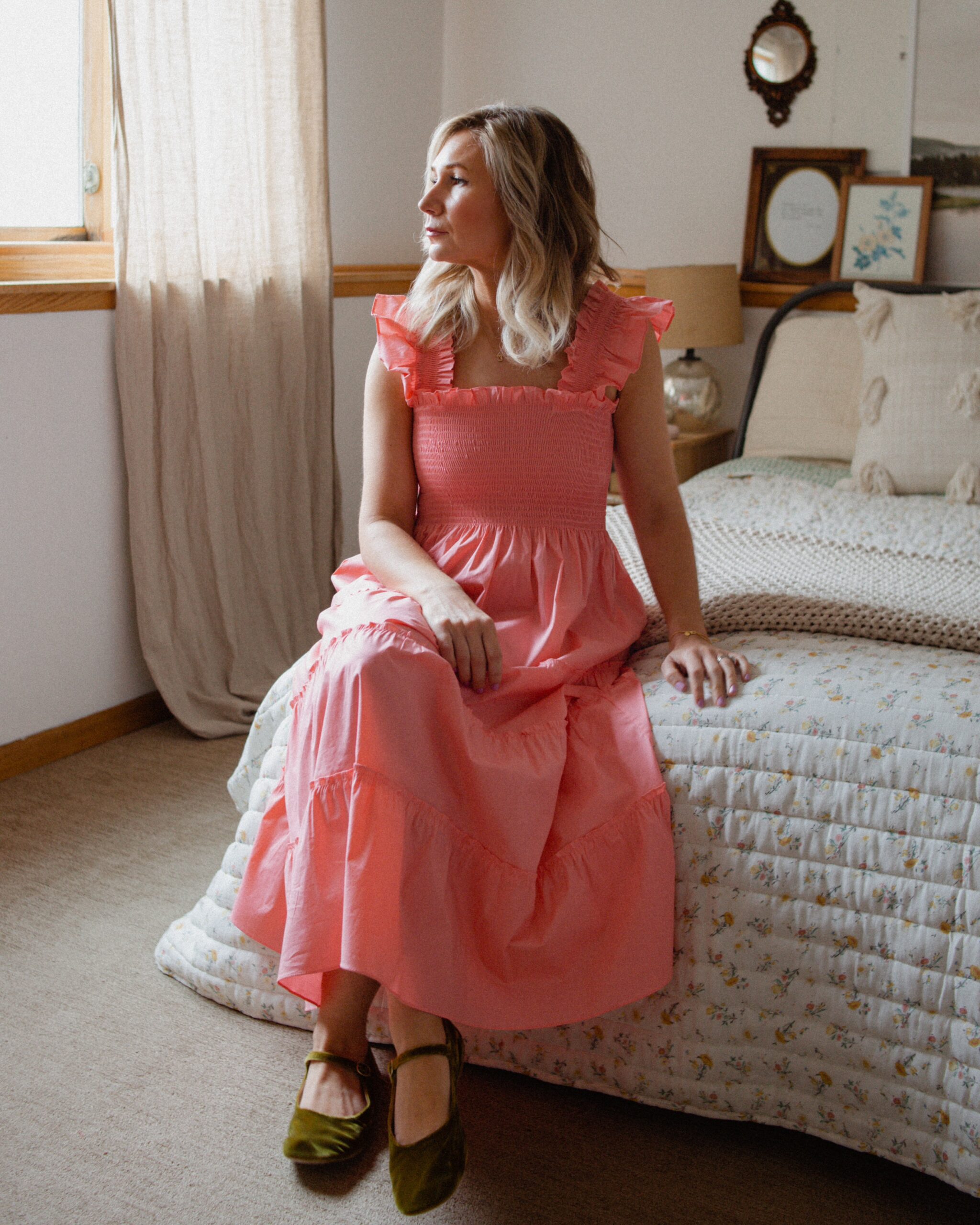 Karin Emily wears a bright pink dress and green velvet mary janes while sitting on a bed sharing her Sephora VIB sale picks