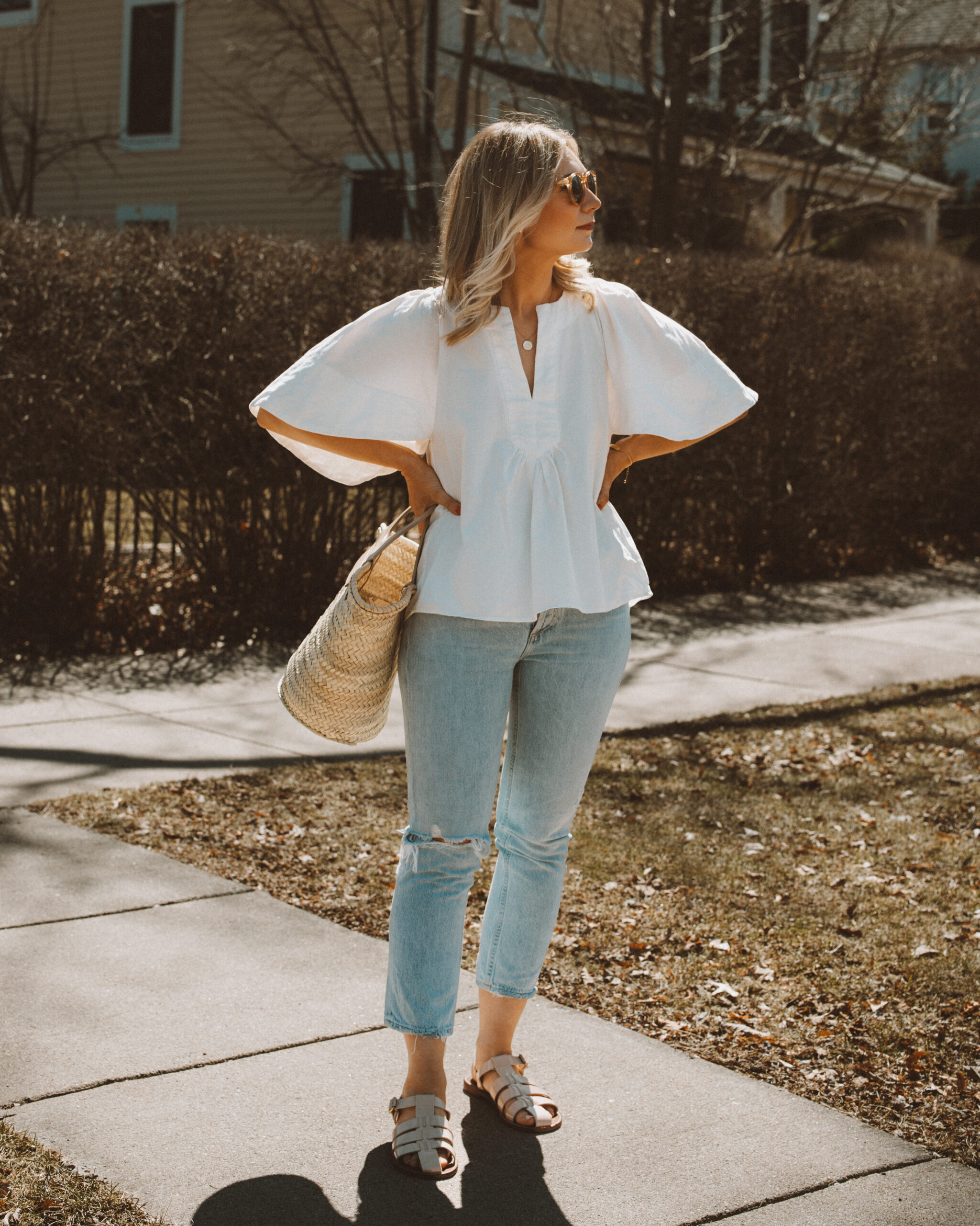 Karin Emily wears a white flutter sleeve blouse with a pair of light blue wash riley jeans from Agolde, a straw Cabas bag from Hereu, fisherman sandals from J. Crew, and clear framed sunglasses