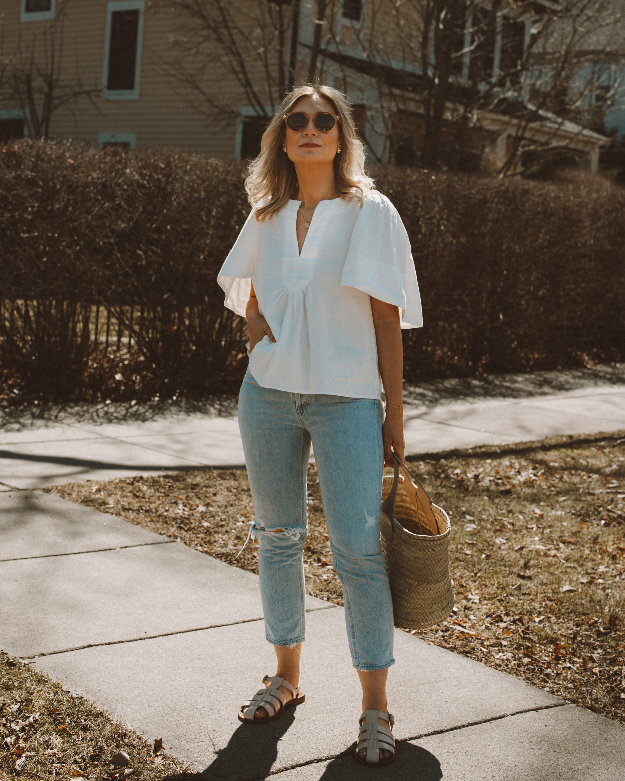Karin Emily wears a spring top with a pair of light blue wash riley jeans from Agolde, a straw Cabas bag from Hereu, fisherman sandals from J. Crew, and clear framed sunglasses