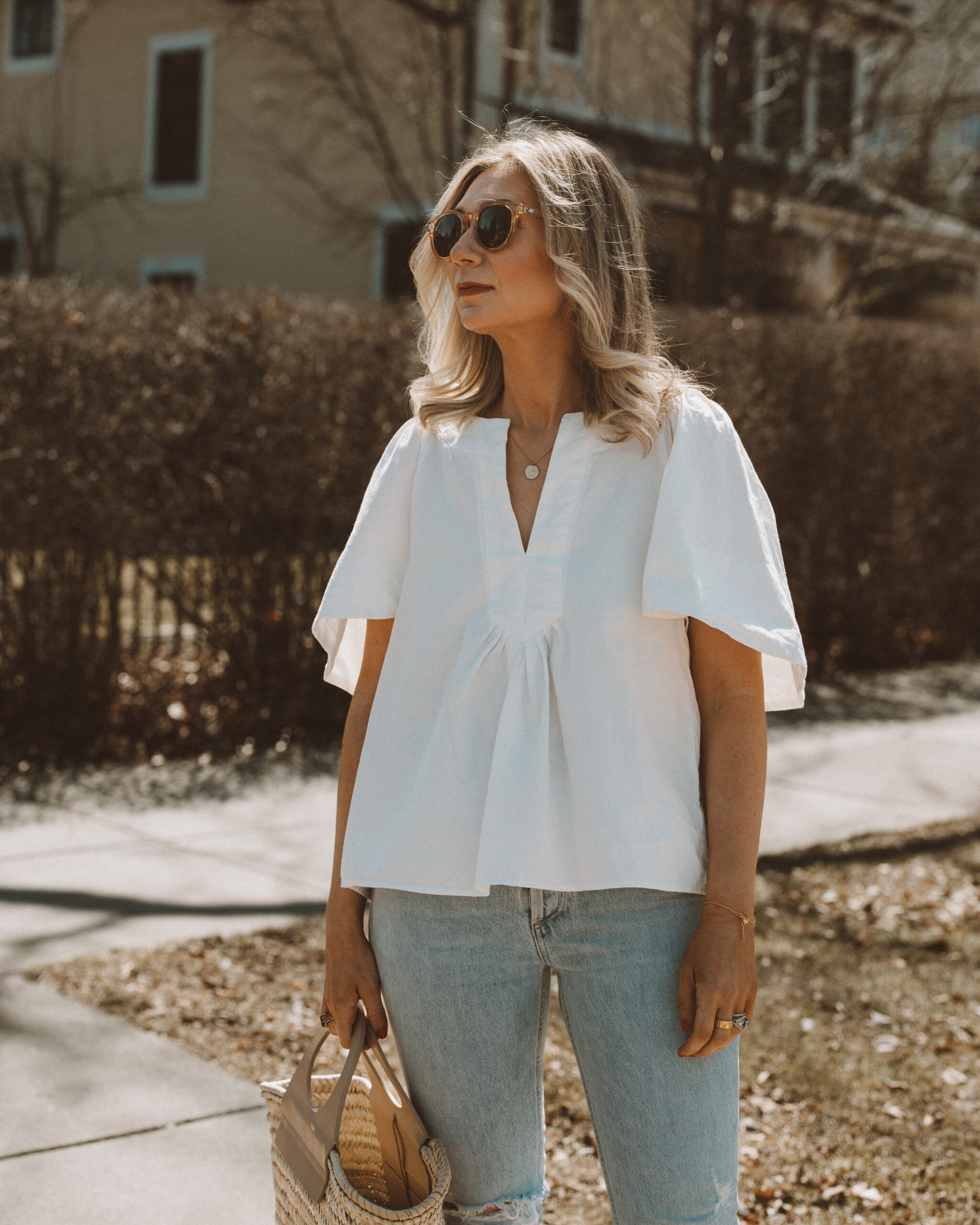 Karin Emily wears a white flutter sleeve blouse with a pair of light blue wash riley jeans from Agolde and clear framed sunglasses