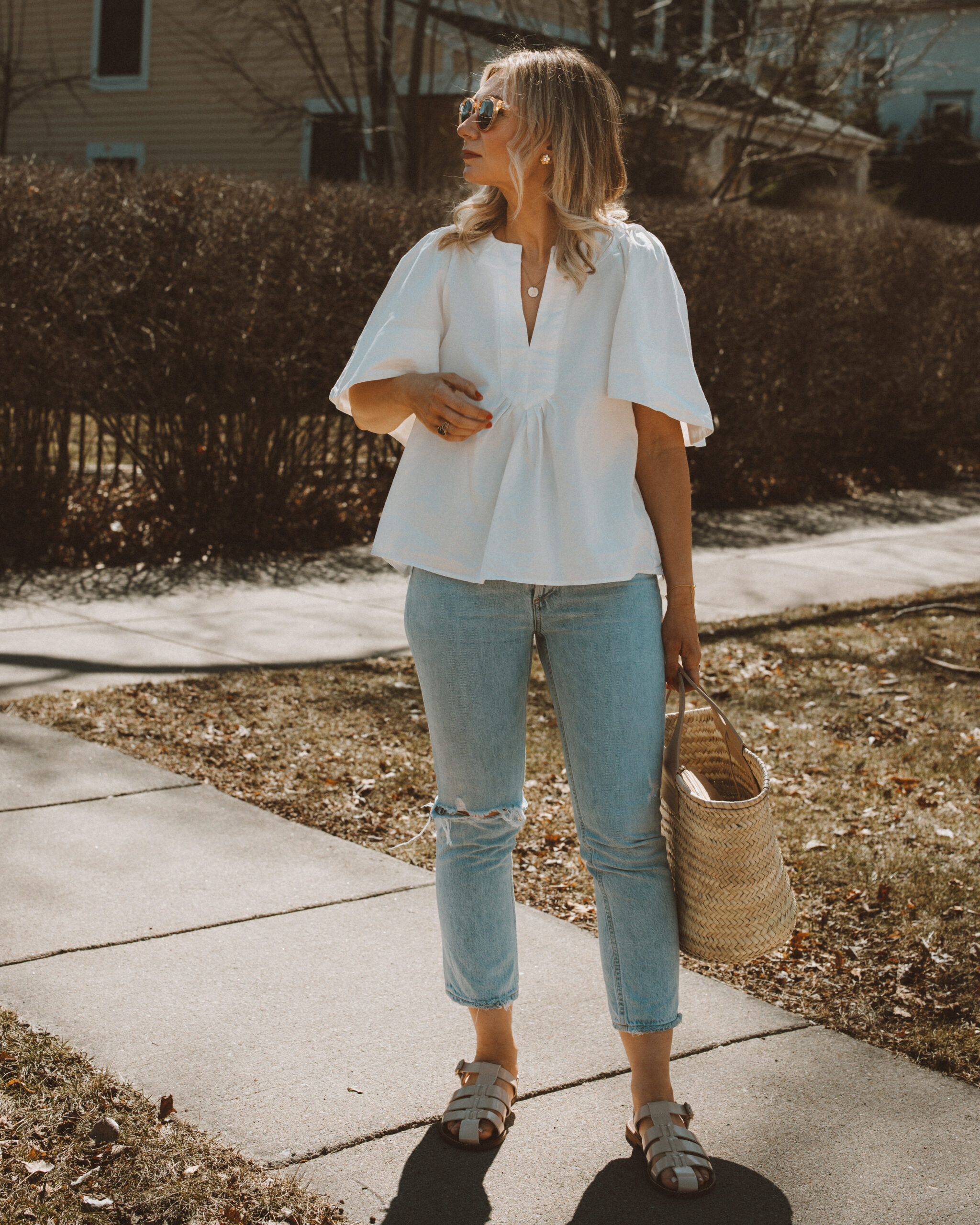 Karin Emily wears a white flutter sleeve blouse with a pair of light blue wash riley jeans from Agolde, a straw Cabas bag from Hereu, fisherman sandals from J. Crew, and clear framed sunglasses