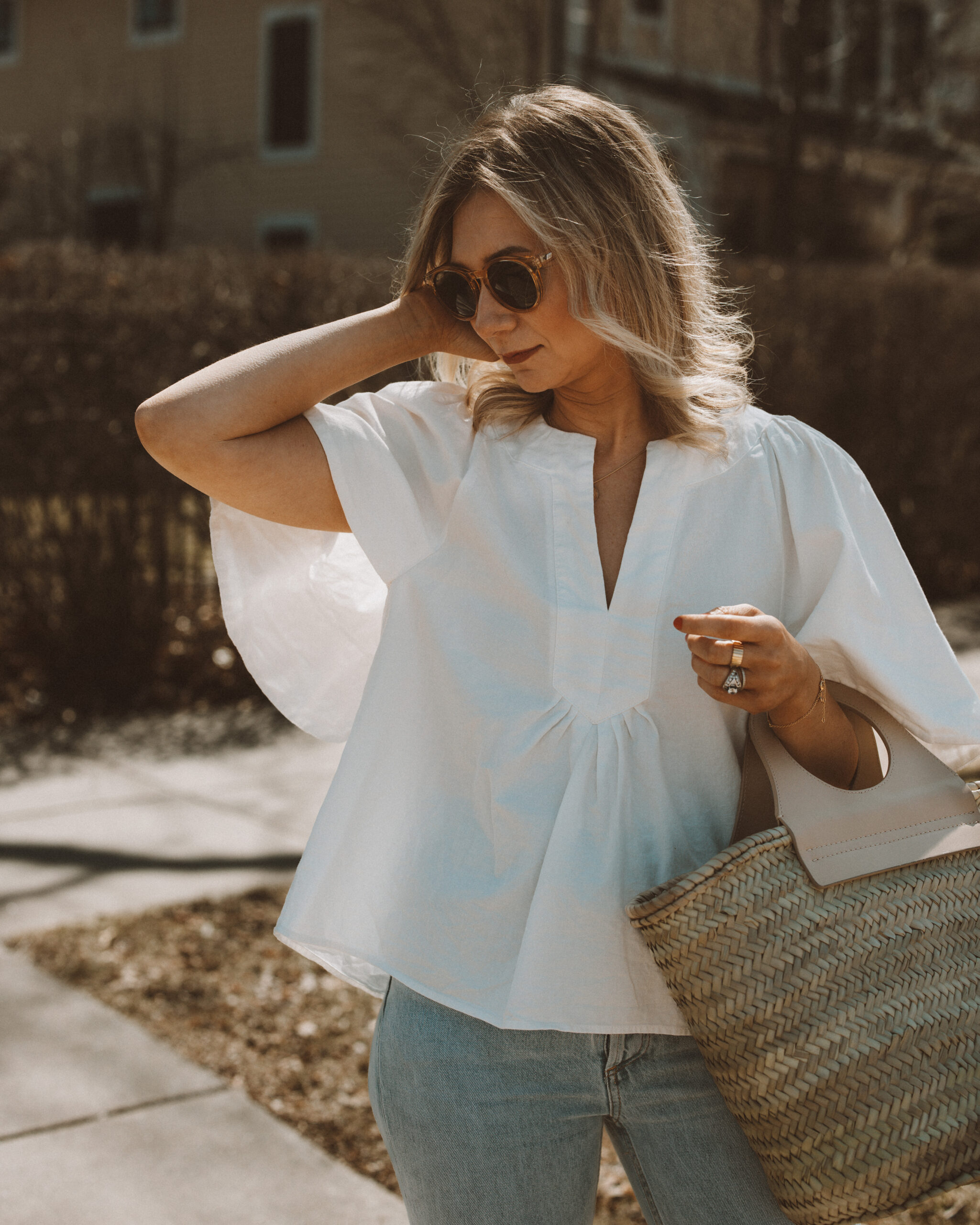 Karin Emily wears a spring top with a pair of light blue wash riley jeans from Agolde, a straw Cabas bag from Hereu, and clear framed sunglasses