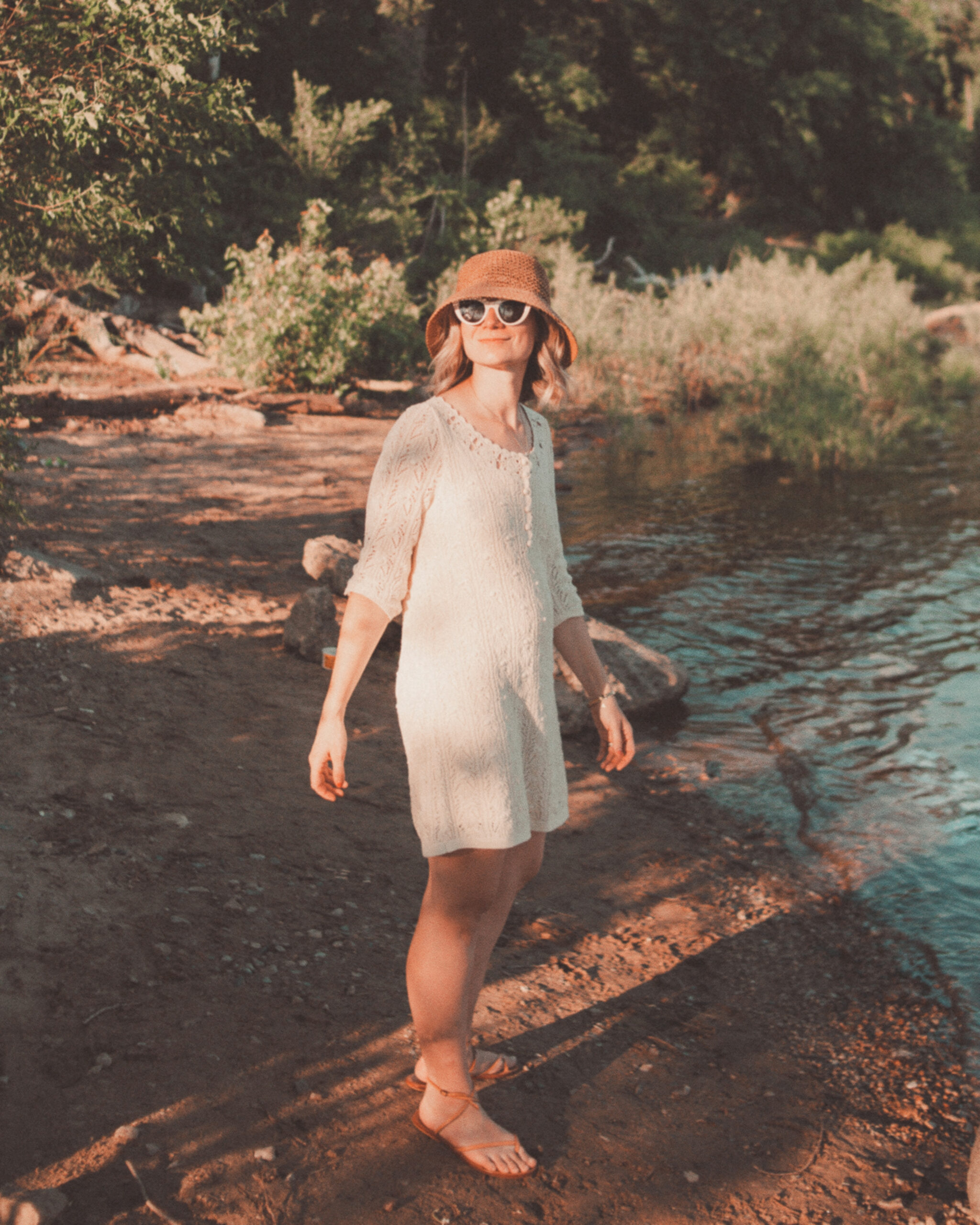 Karin Emily wears a little crochet dress from Sezane and a straw bucket hat from Vitamin A in front of a riverfront