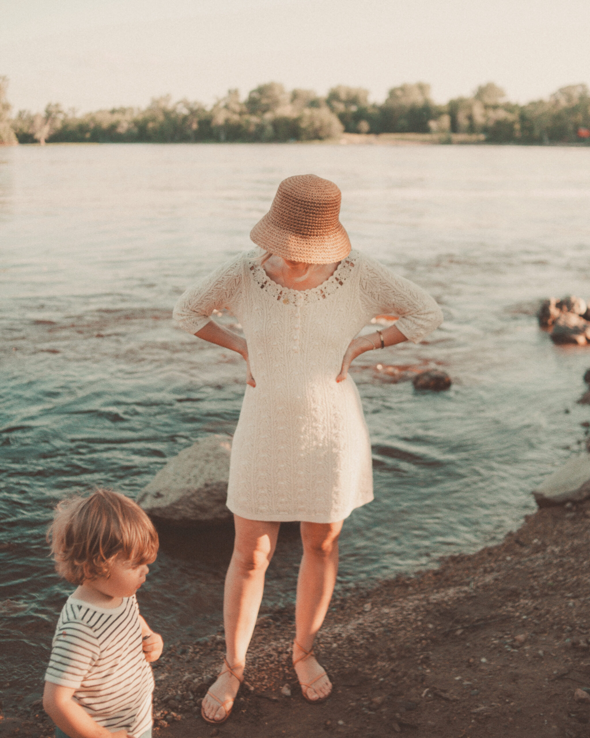 Karin Emily wears a little crochet dress from Sezane and a straw bucket hat from Vitamin A in front of a riverfront
