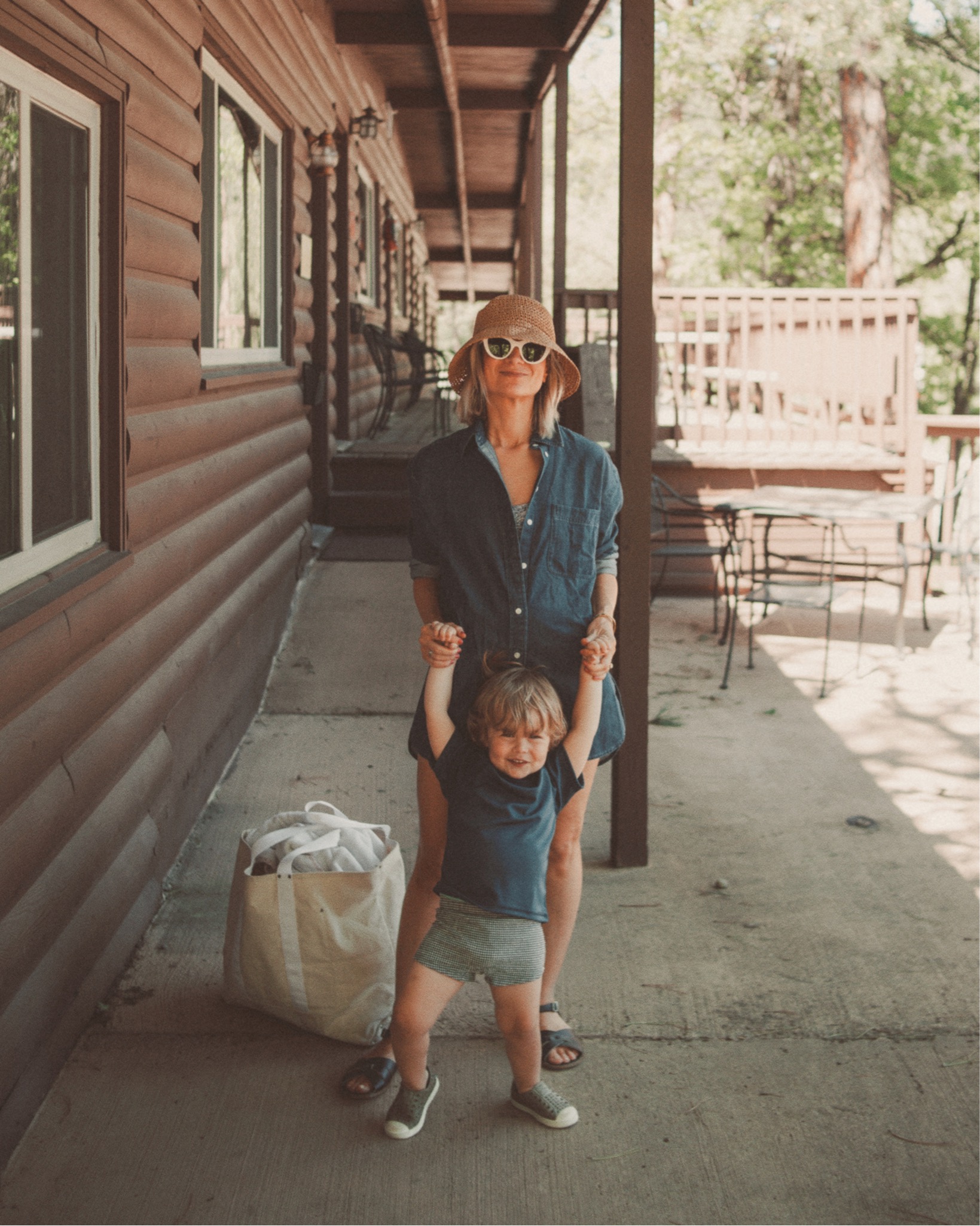 Karin Emily wears a blue long sleeve button down swimsuit coverup over a blue and white floral one piece swimsuit and a pair of black saltwater sandals with her son in a navy blue rashguard and gingham european style swim trunks